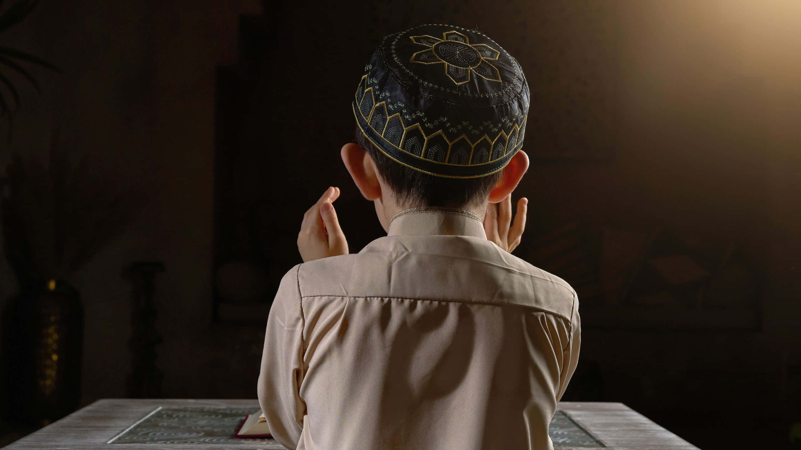 Praying 桌布 壁紙 — 一個戴著圖案帽的男孩在昏暗的房間裡，被溫暖的背光包圍，舉起雙手祈禱。