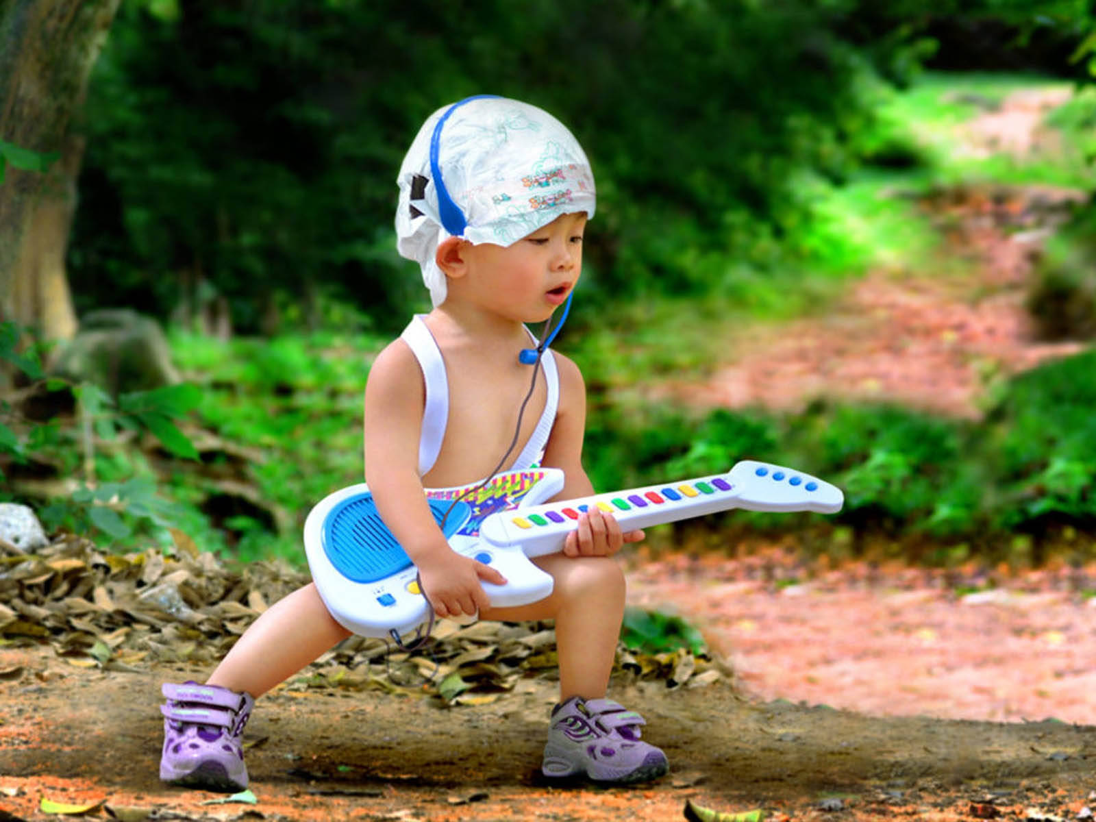 Funny Baby 桌布 壁紙 — 一個戴著塑膠頭盔和紫色鞋的幼兒，在鋪滿樹葉的森林小徑上蹲著，彈奏色彩繽紛的玩具吉他。
