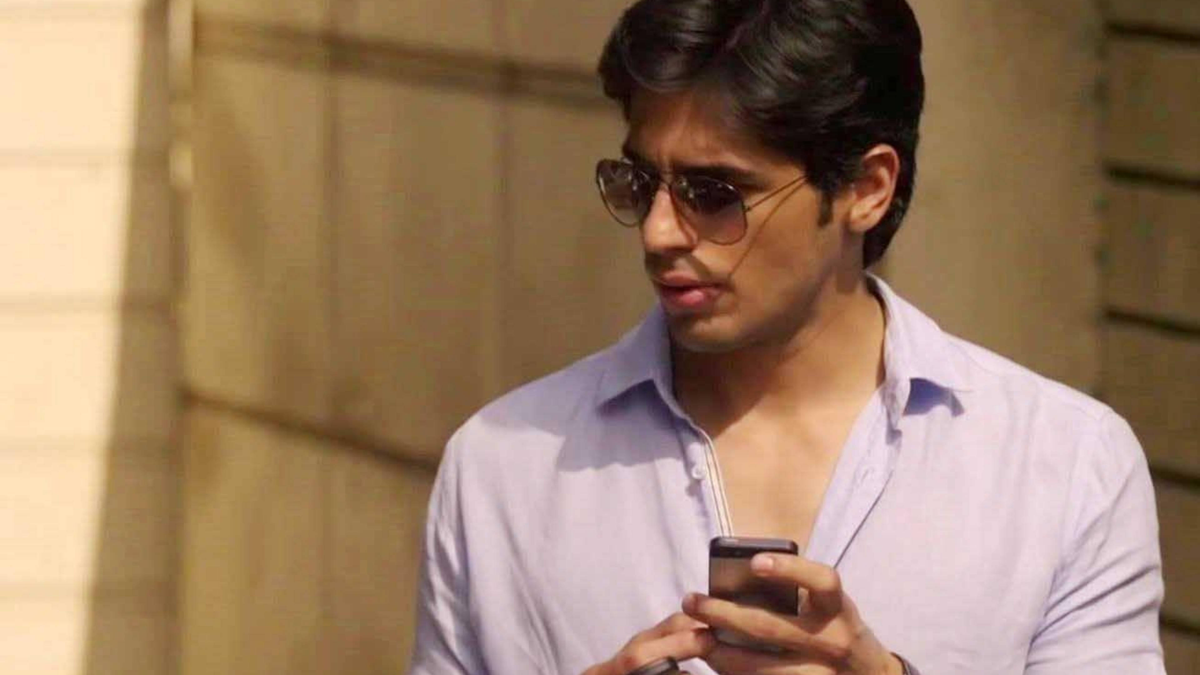 Sidharth Malhotra 桌布 壁紙 — 一個戴著太陽眼鏡、身穿淺色襯衫的男人，一邊走過陽光普照的建築物外牆，一邊專注地看著手機。