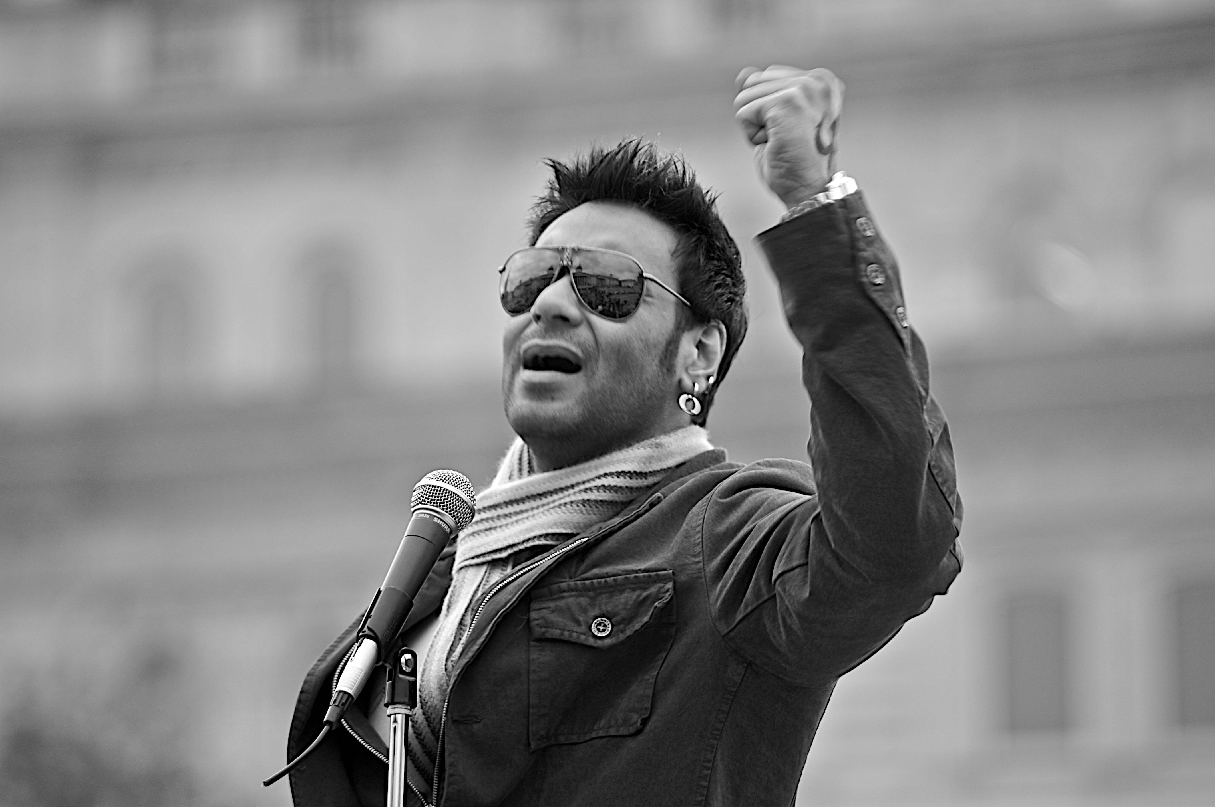 ajay devgn 桌布 壁紙 — 一個戴著太陽眼鏡和圍巾的男人，握緊拳頭高舉空中，熱情地對著麥克風說話。