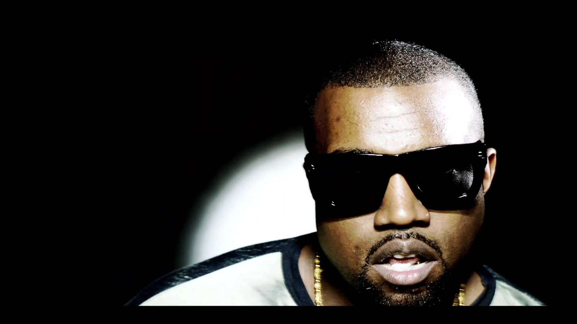 Kanye 桌布 壁紙 — 一個戴著太陽眼鏡和金色鏈條的男人，在明亮的聚光燈下，對著純黑色背景強烈凝視。