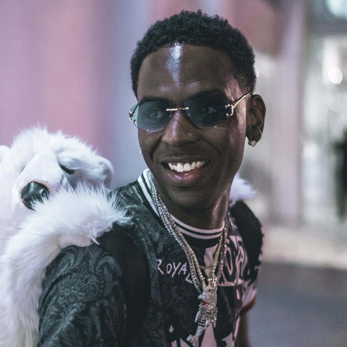 Young Dolph 桌布 壁紙 — 一個戴著太陽眼鏡和閃亮珠寶的微笑男子，在模糊的城市背景下抱著一隻白色毛茸茸的狗。