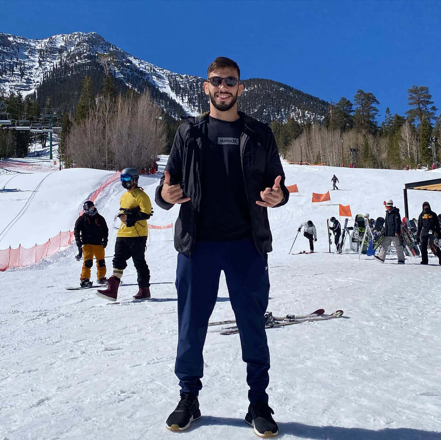 Snowy Mountain 桌布 壁紙 — 一個戴著太陽眼鏡微笑的男人在陽光明媚的雪坡上豎起兩個大拇指，背景有山脈和其他滑雪者。