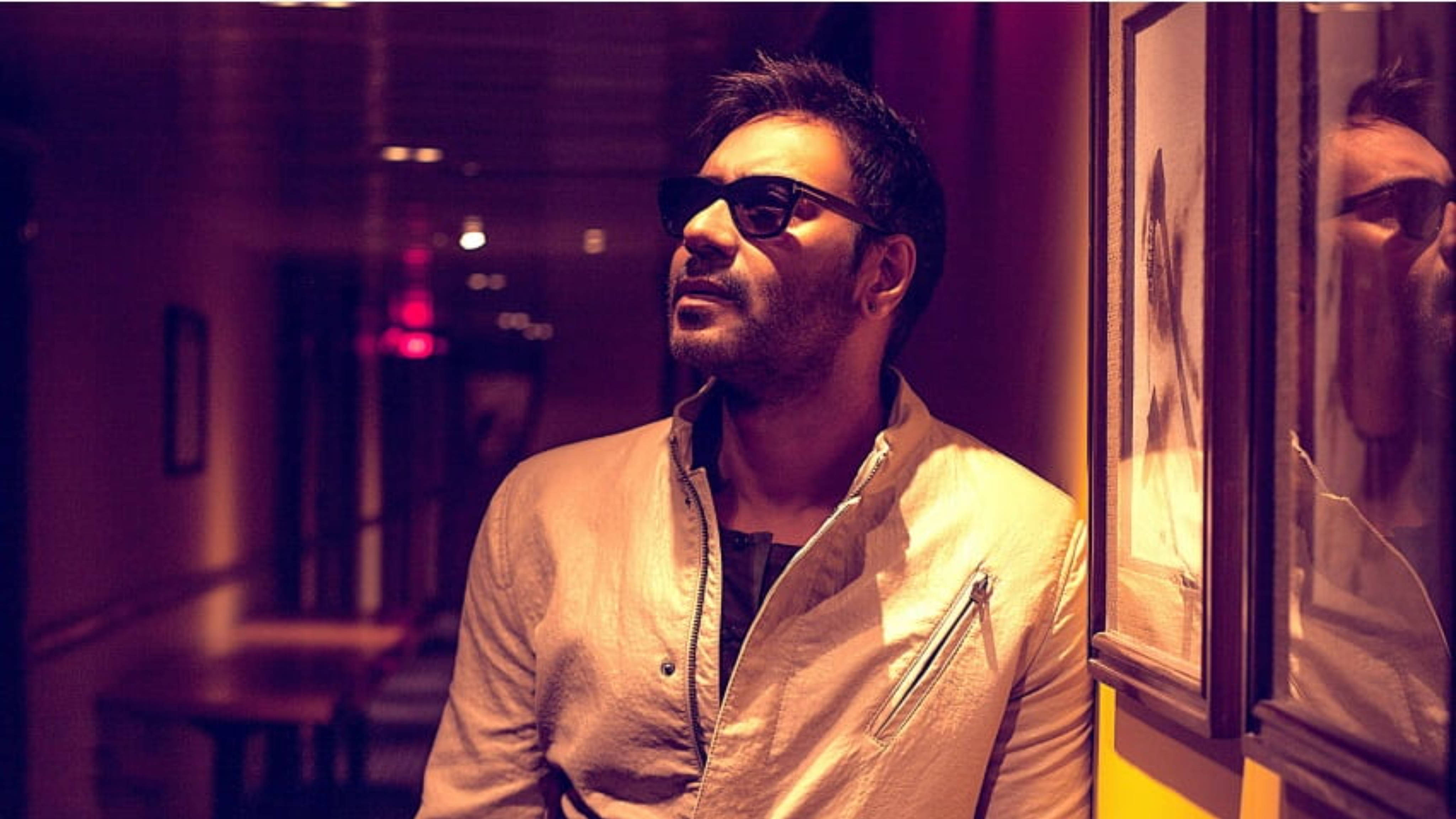 ajay devgn 桌布 壁紙 — 一個戴著太陽眼鏡的男人靠在溫暖照明的走廊牆上，牆上有裱框藝術品和柔和環境光。