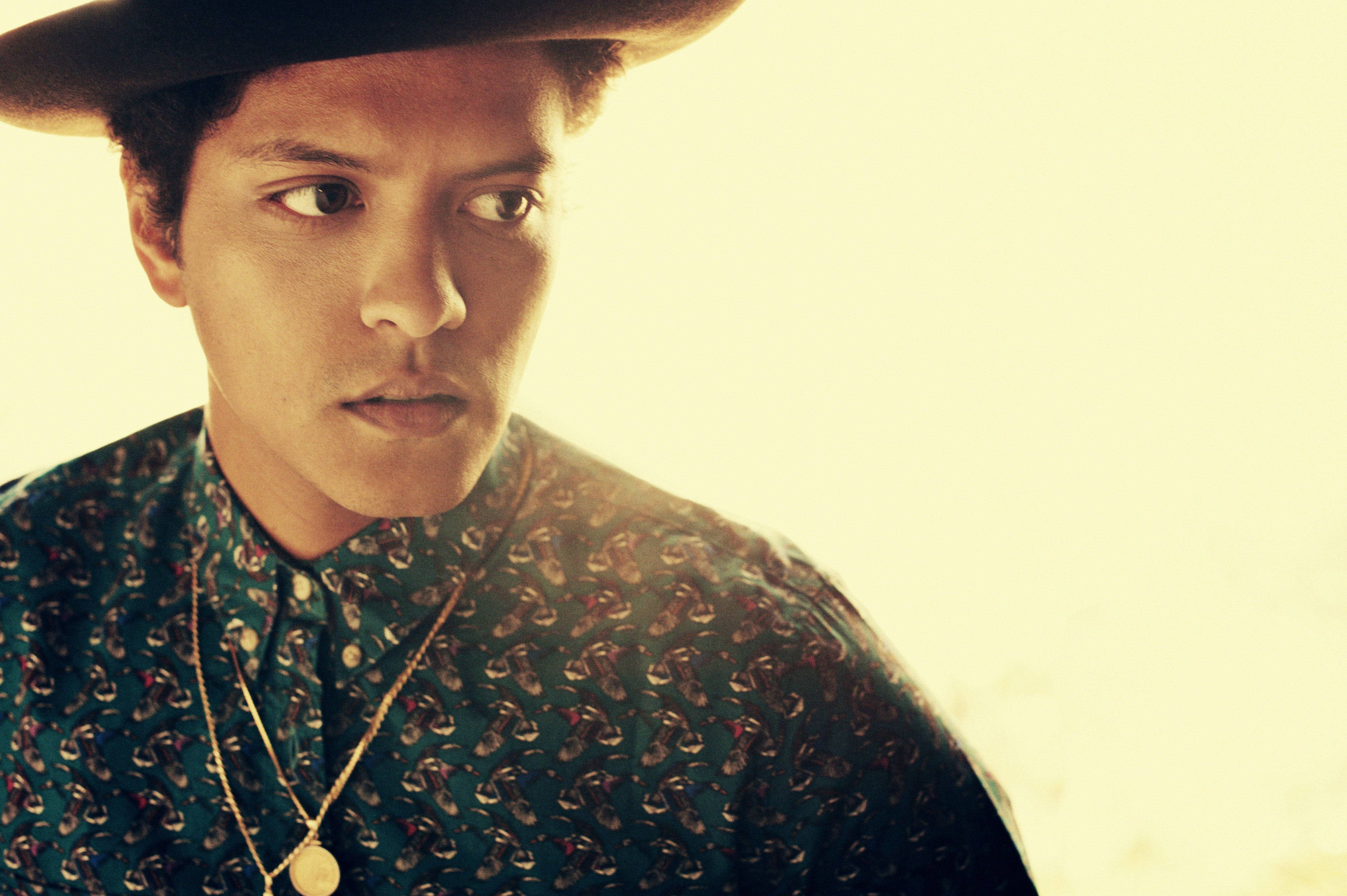 Bruno Mars 桌布 壁紙 — 一個戴著寬邊帽和圖案襯衫的男人，在溫暖發光的背景下若有所思地凝視著。