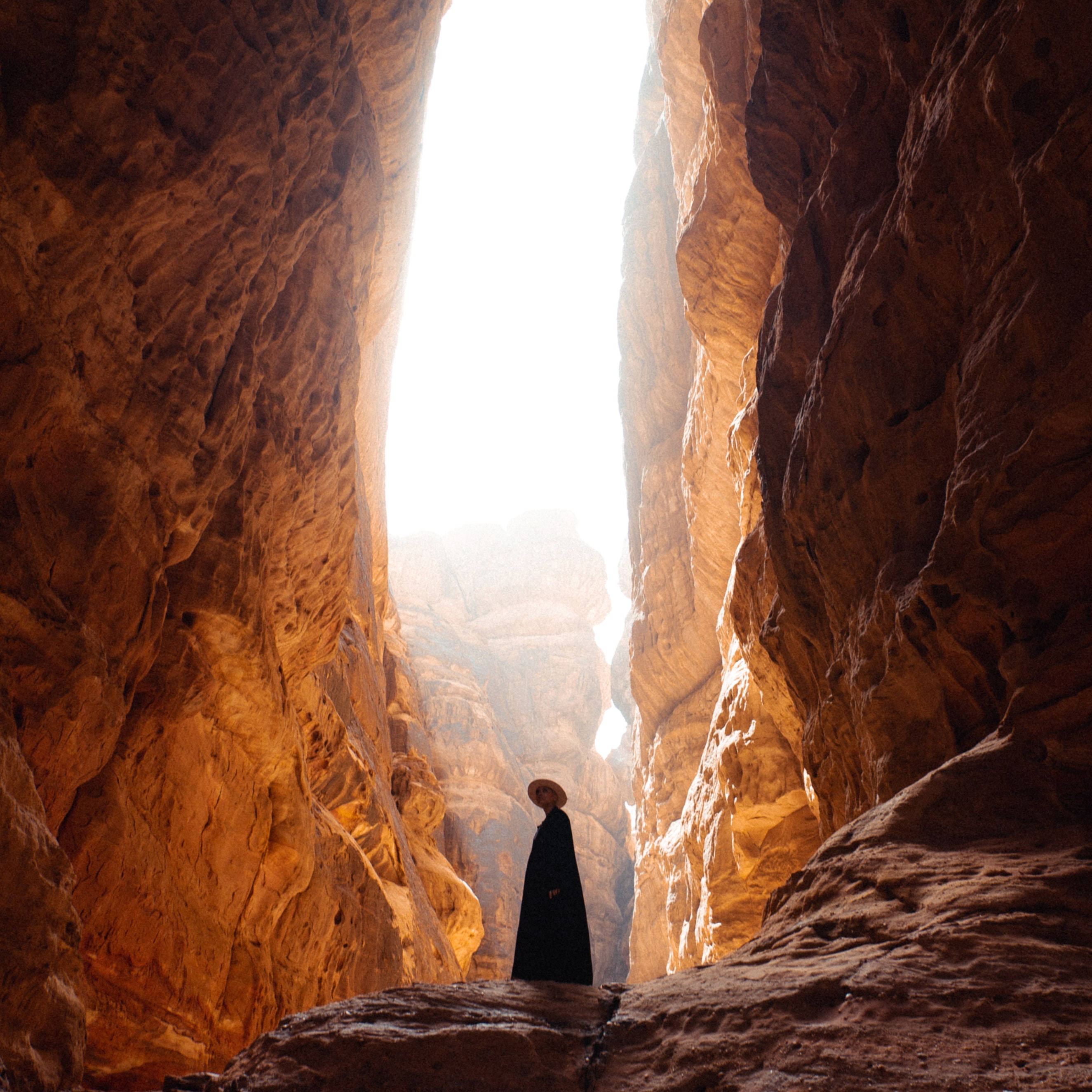 Saudi 桌布 壁紙 — 一個戴著寬邊帽的孤獨身影，在狹窄峽谷的開口處形成剪影，沐浴在透過高聳紅色岩壁過濾的金色陽光中。