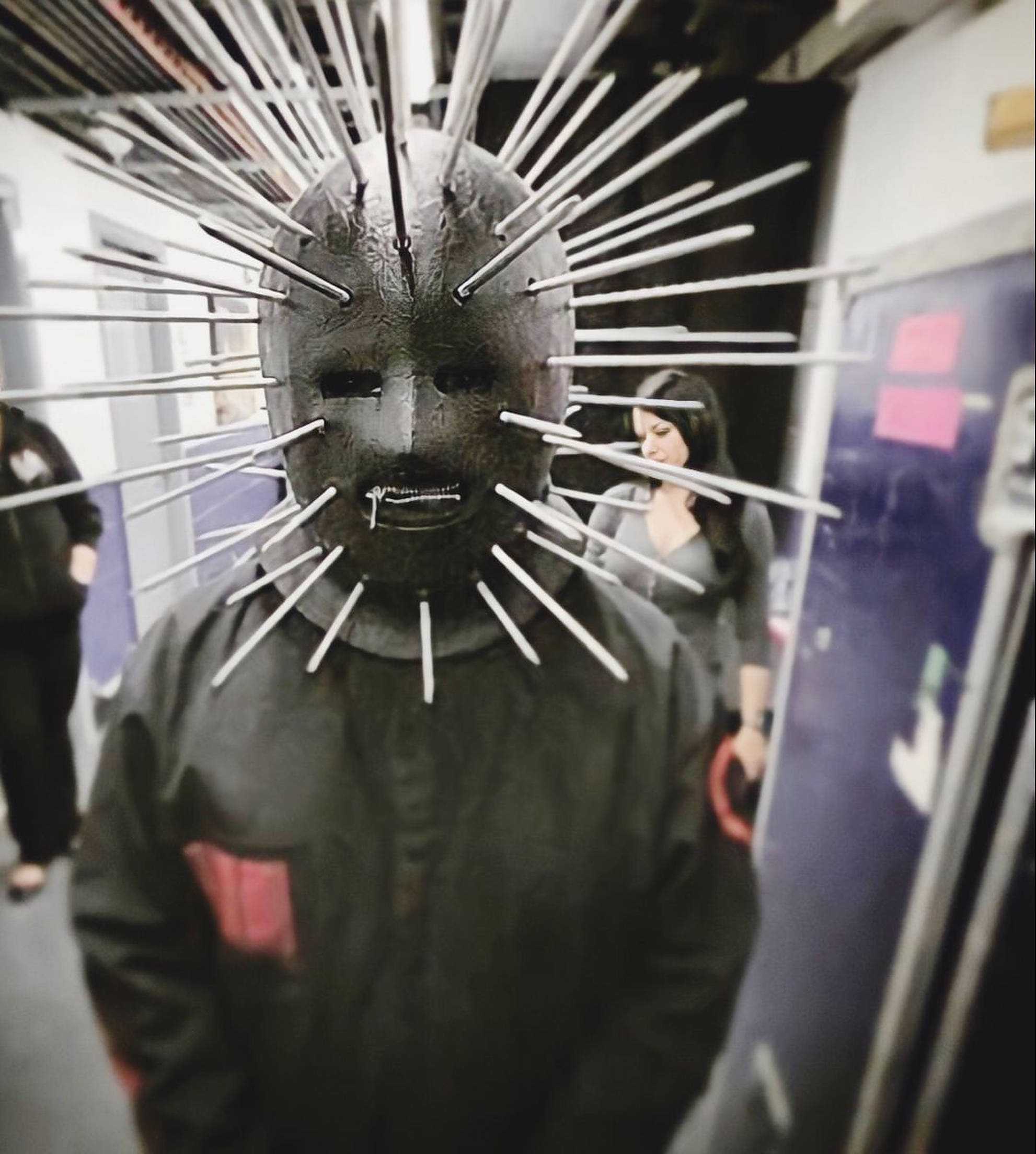 craig jones 桌布 壁紙 — 一個戴著尖刺Pinhead面具的人站在地鐵車廂內，背景中一名女子走過。