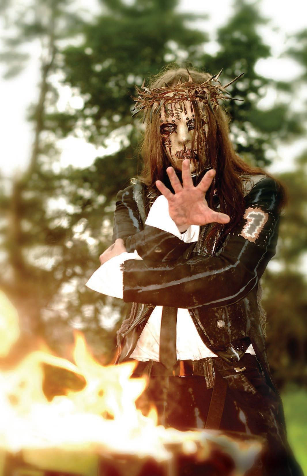Joey Jordison 桌布 壁紙 — 一個戴著尖刺王冠、臉部塗繪的人站在森林中熊熊燃燒的火堆前，舉起手做出停止手勢。