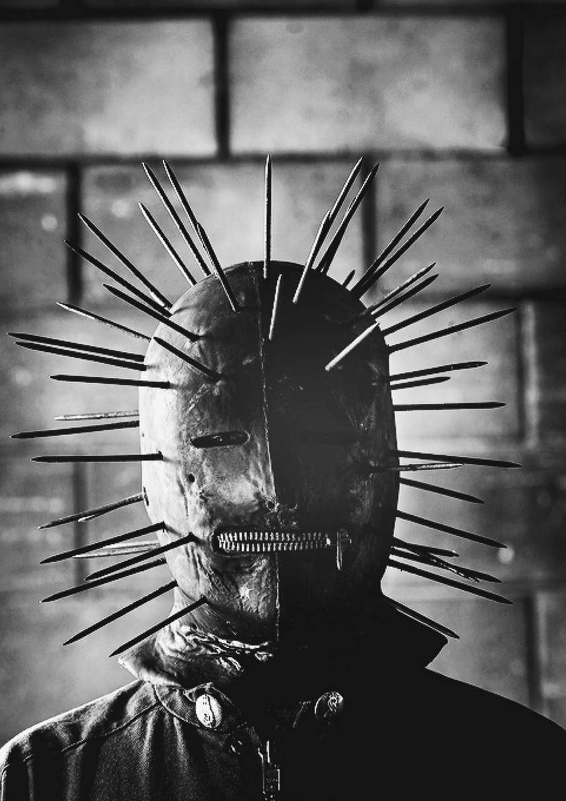 craig jones 桌布 壁紙 — 一個戴著尖刺面具和拉鍊嘴的威脅人物，在鮮明的黑白背景下靠著磚牆站立。