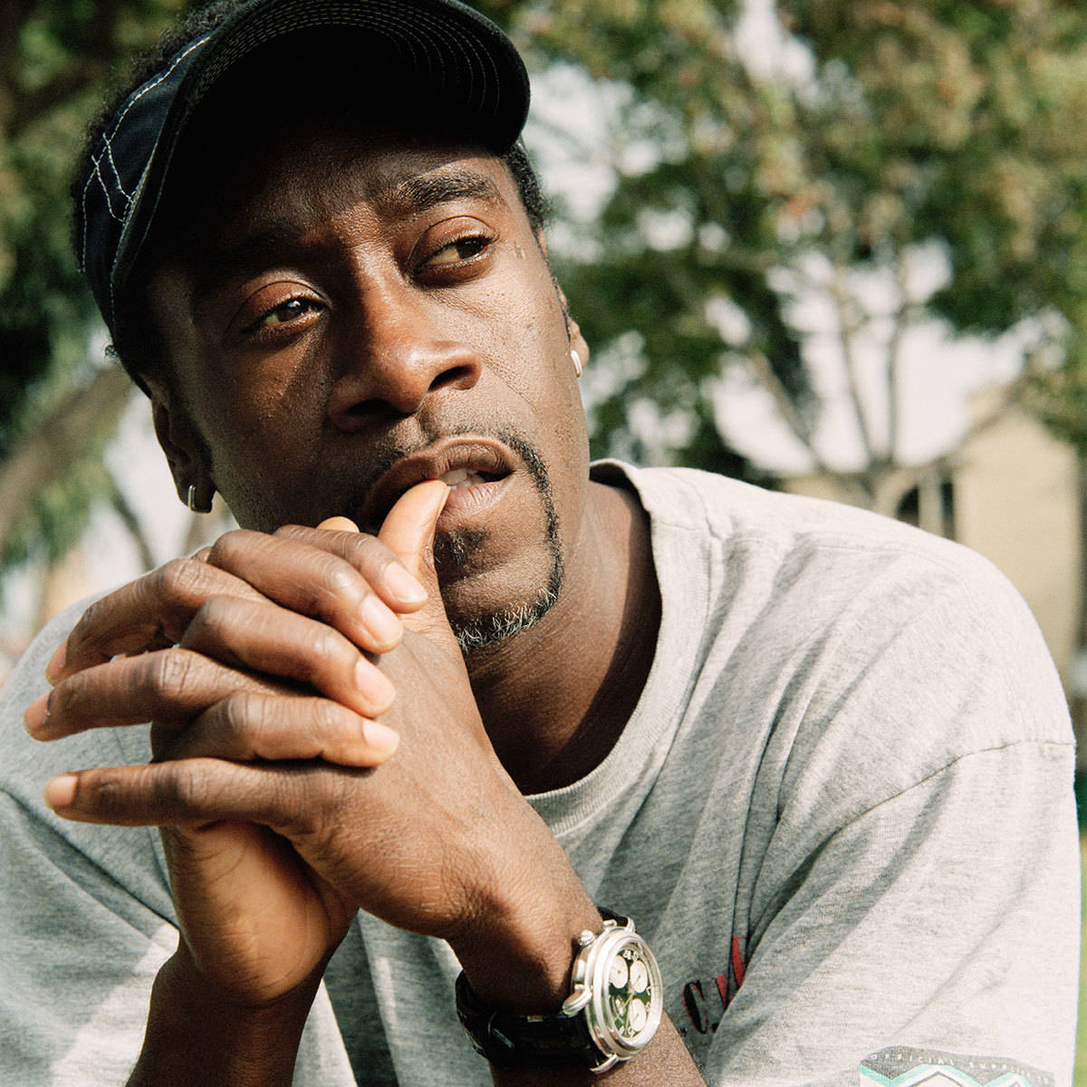 Don Cheadle 桌布 壁紙 — 一個戴著帽子的沉思男子，在陽光明媚的戶外背景下，將下巴擱在緊握的雙手上。