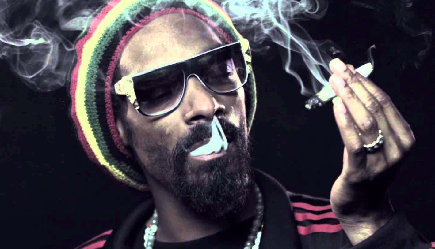 Young Snoop Dogg 桌布 壁紙 — 一個戴著彩色毛帽和太陽眼鏡的男人，在黑暗背景下手持點燃的香煙呼出煙霧。