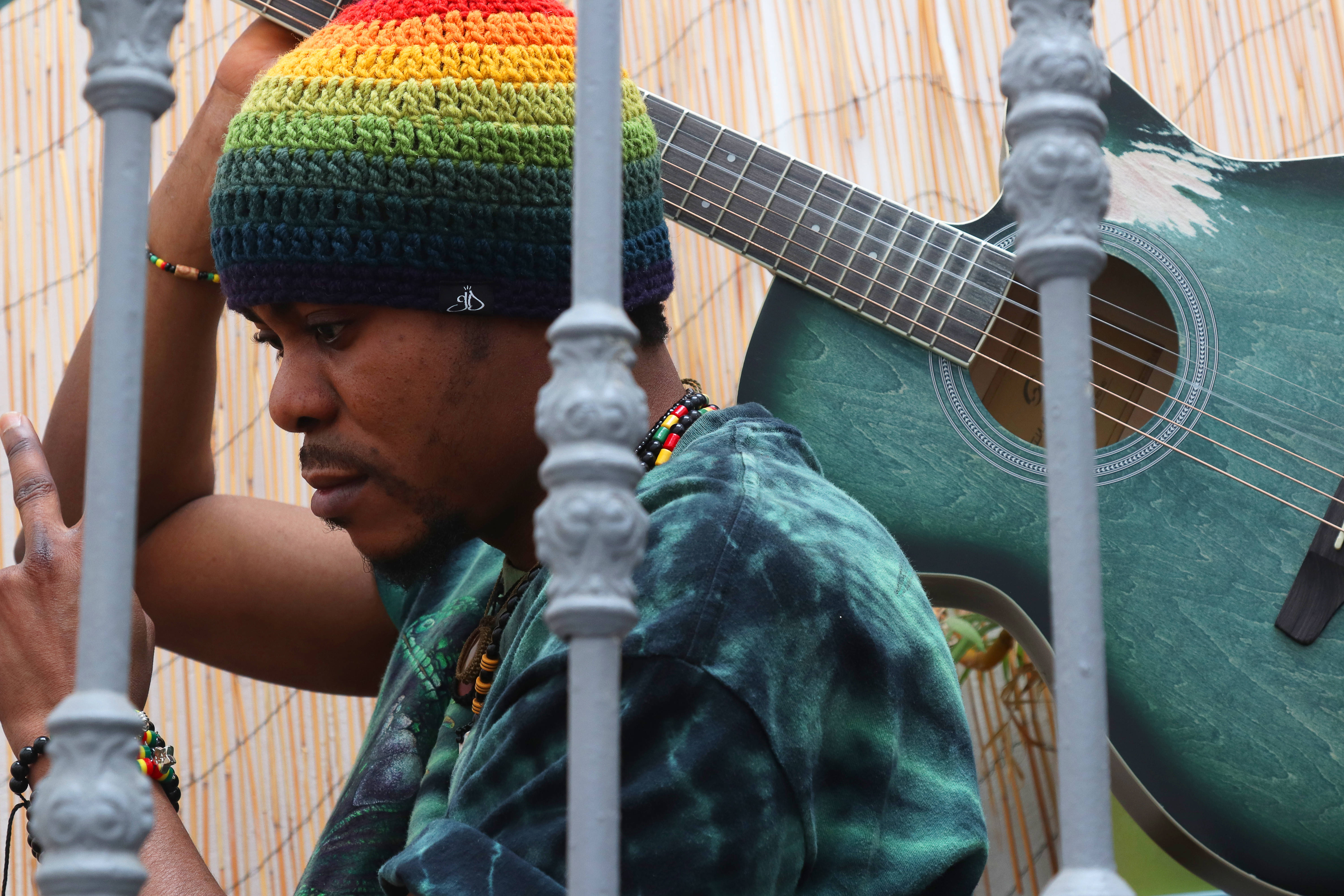 Reggae 桌布 壁紙 — 一個戴著彩虹針織帽和紮染襯衫的男人靠在華麗的灰色欄杆旁，旁邊有一把綠色木吉他。