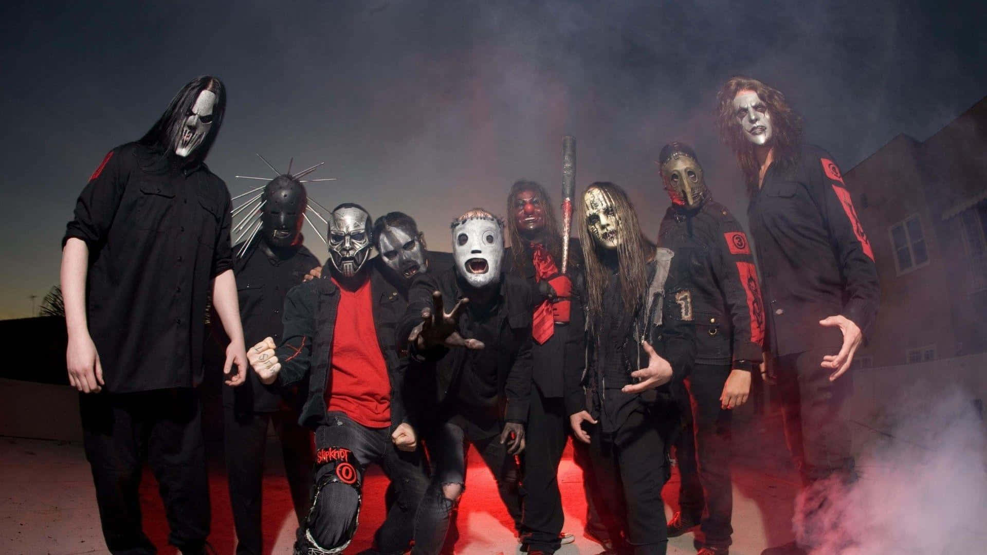 Slipknot 桌布 壁紙 — 一個戴著怪誕面具、身穿黑色服裝的樂團在煙霧瀰漫的暮色中，在紅色燈光下戲劇性地擺姿勢。