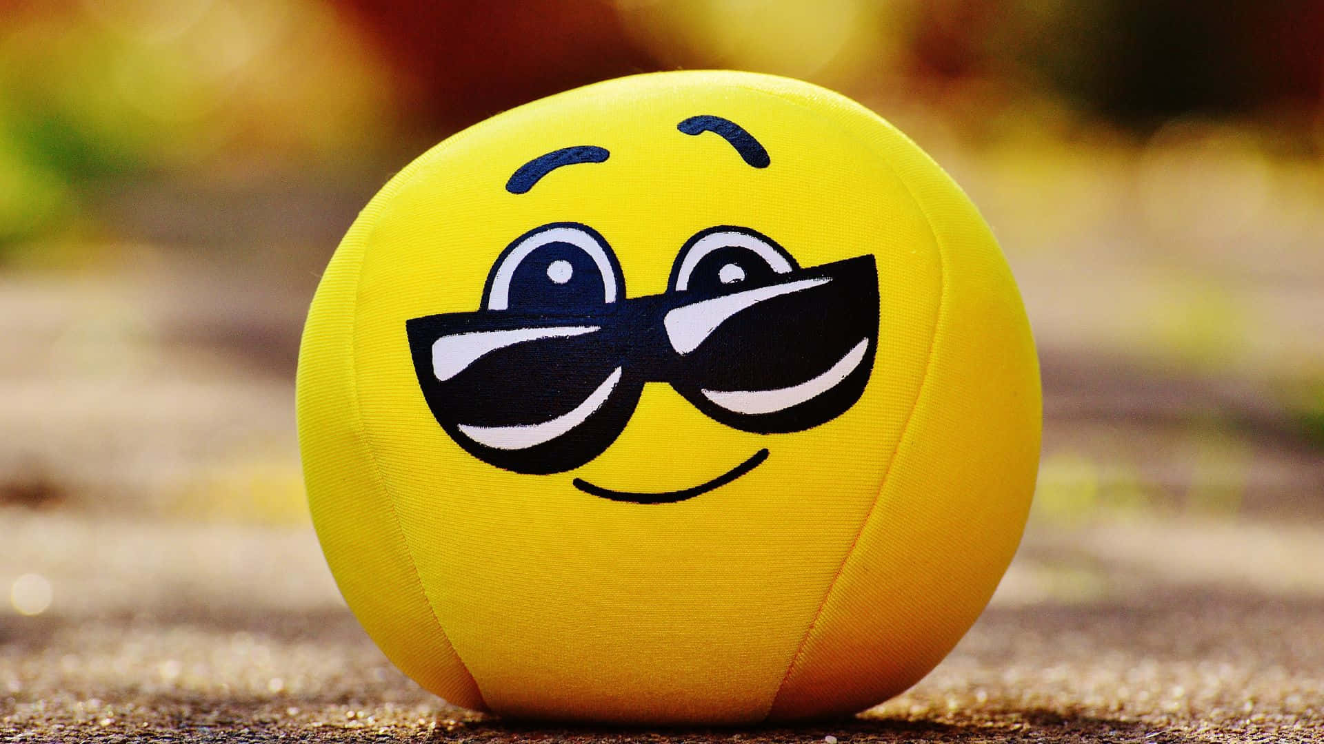 Smiley 桌布 壁紙 — 一個戴著時尚太陽眼鏡的開心黃色表情符號球，坐在沙地上，背後有模糊的秋葉。