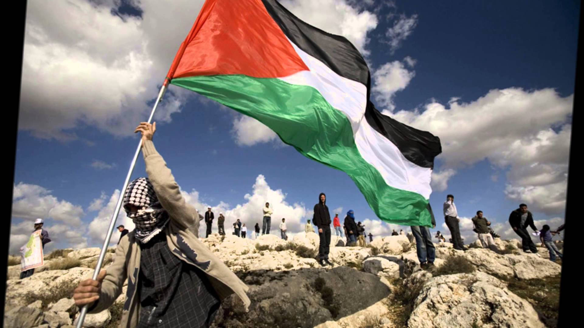 Palestine 桌布 壁紙 — 一個戴著格子頭巾的人在岩石山丘上，在晴朗藍天和零星白雲下舉起巴勒斯坦旗。