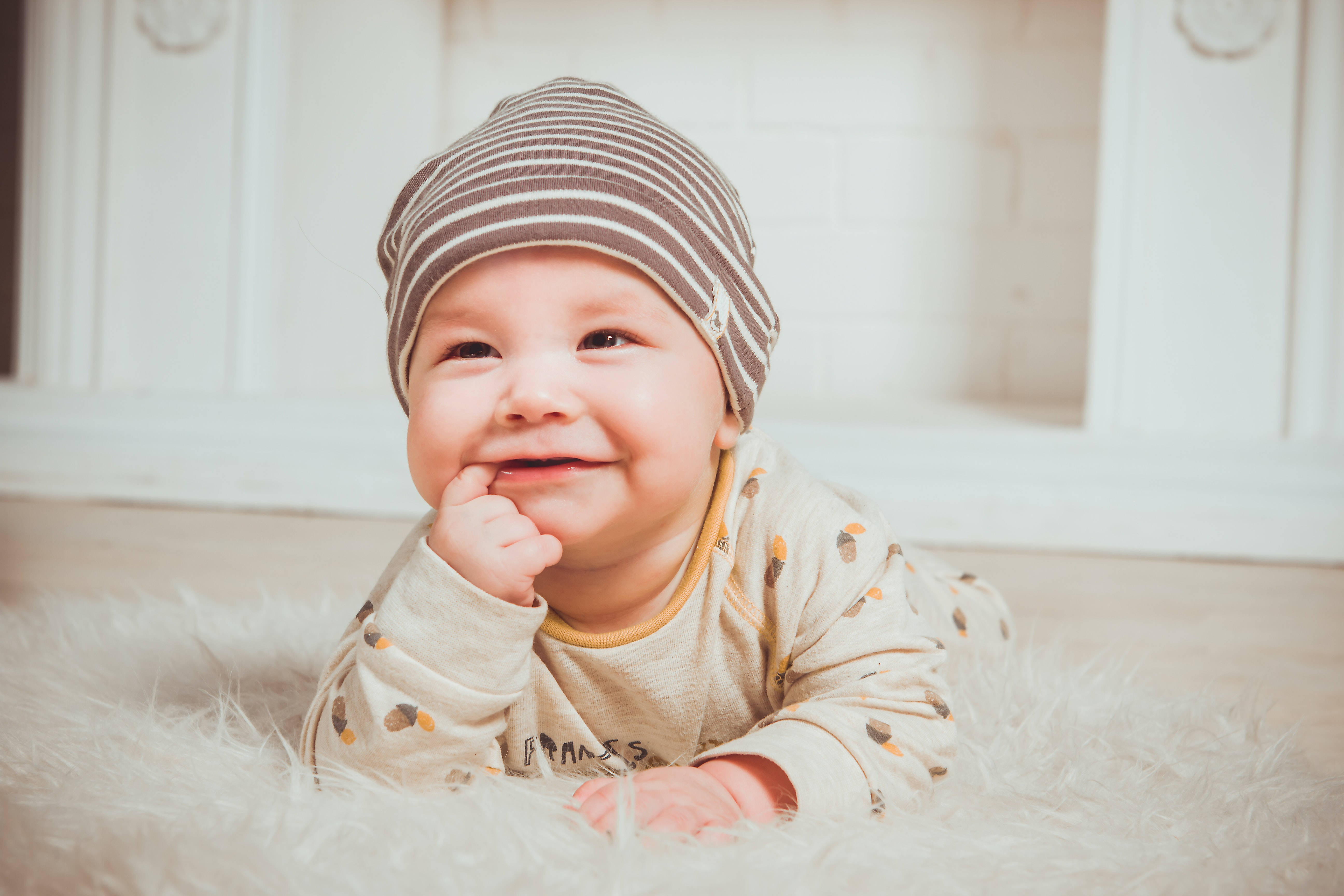 Baby Boy 桌布 壁紙 — 一個戴著條紋帽子的微笑嬰兒躺在室內柔軟的白色地毯上，好奇地看著相機。