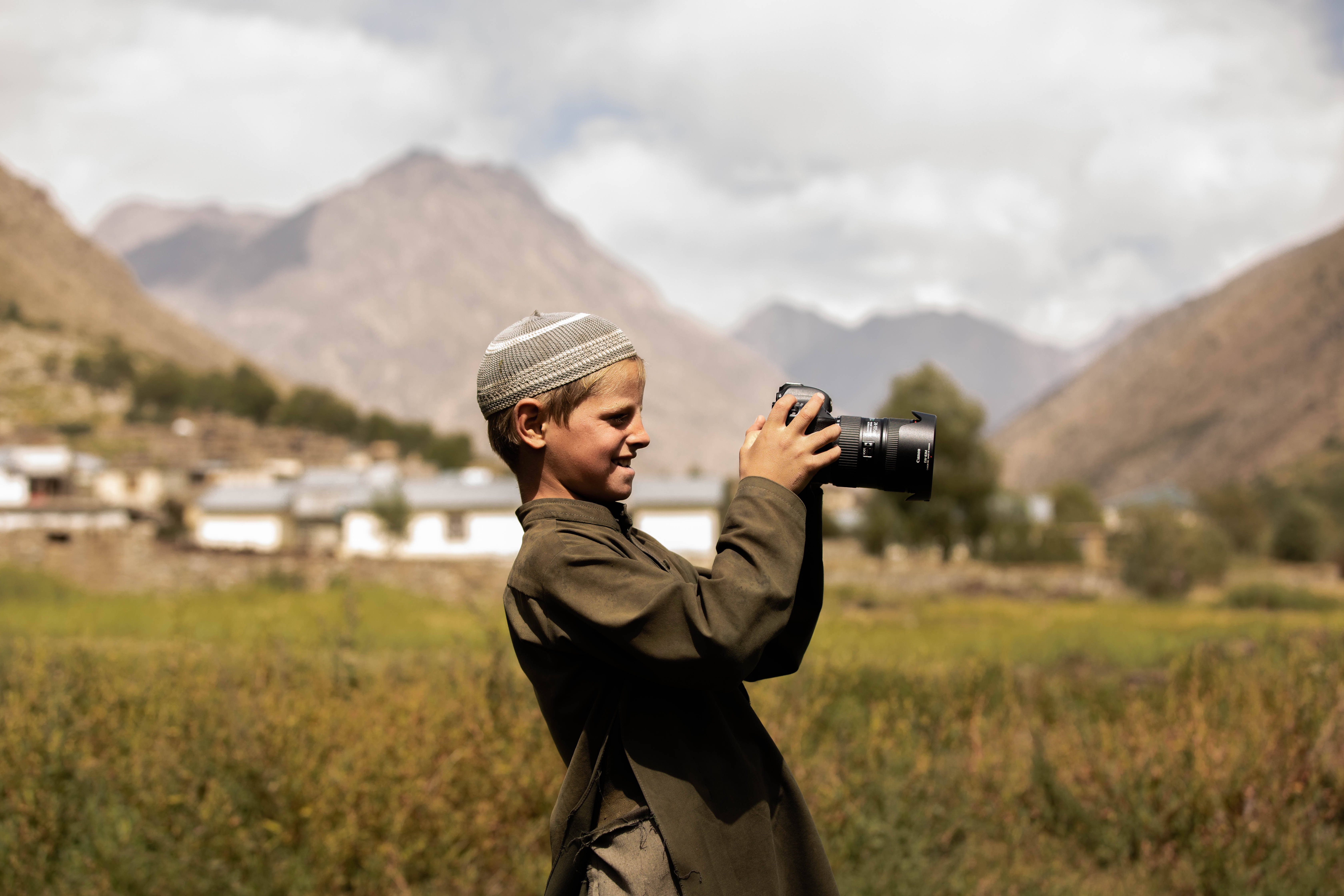 Afghanistan 桌布 壁紙 — 一個戴著條紋帽的男孩微笑著，在山谷中手持相機，背景有石屋和綠色田野。