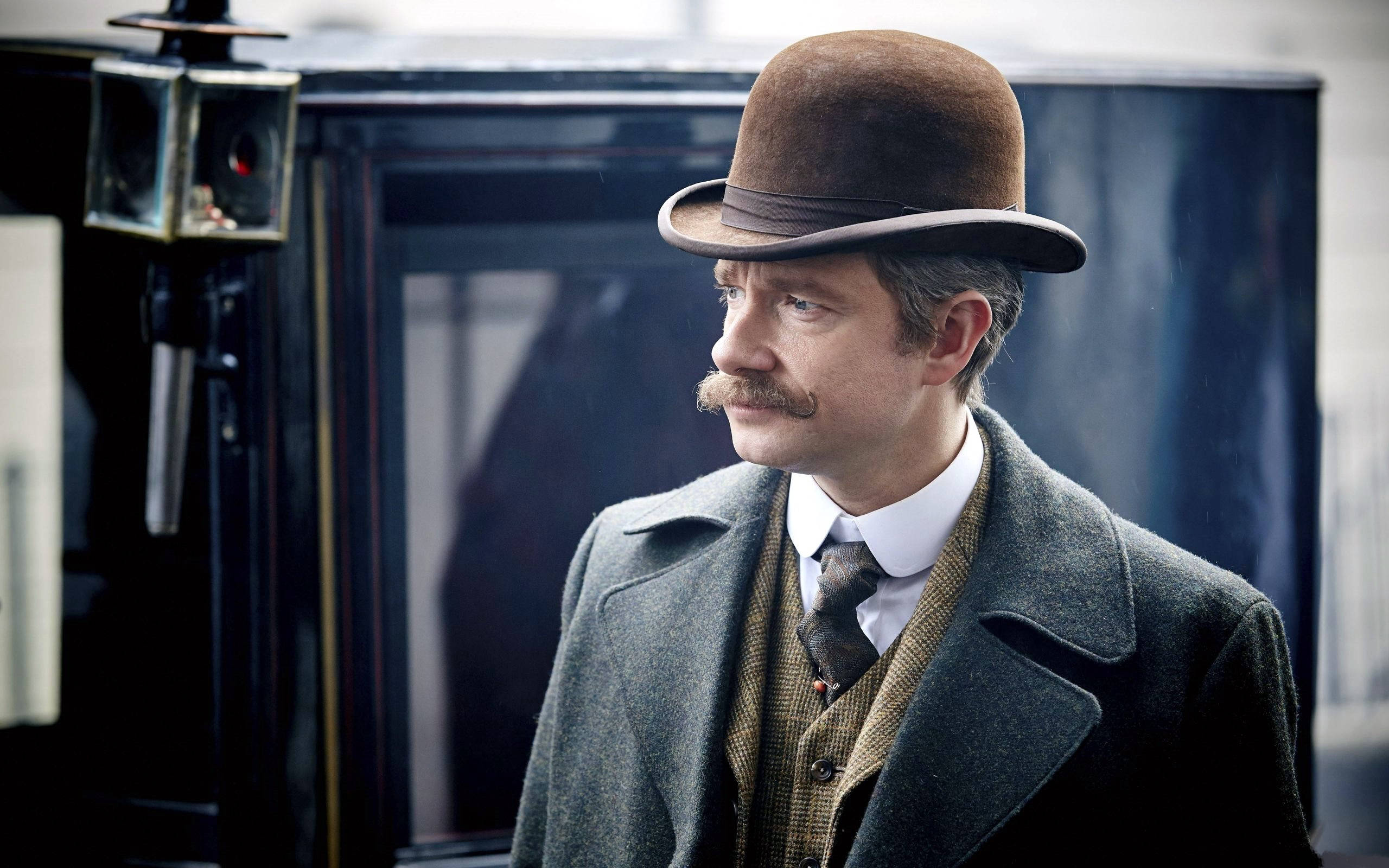 Martin Freeman 桌布 壁紙 — 一個戴著棕色圓頂禮帽和灰色外套的男人，表情嚴肅地站在一輛復古馬車旁邊。