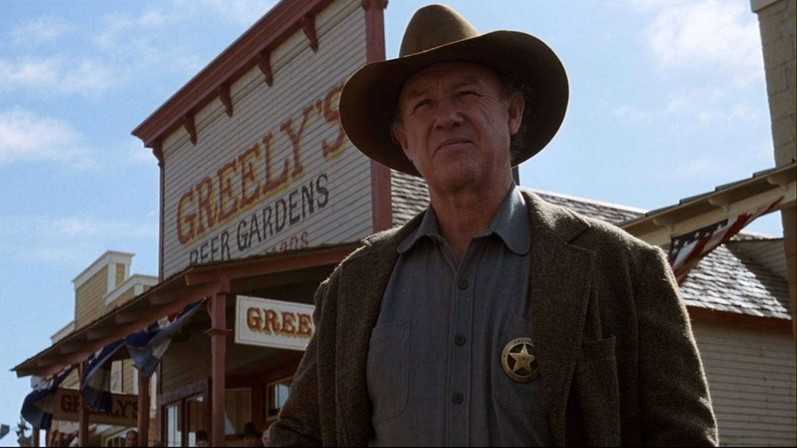 Gene Hackman 桌布 壁紙 — 一個戴著棕色帽子和星形徽章的嚴肅牛仔警長，站在名為 Greely's Beer Gardens 的沙龍前，背景是明亮的藍天。