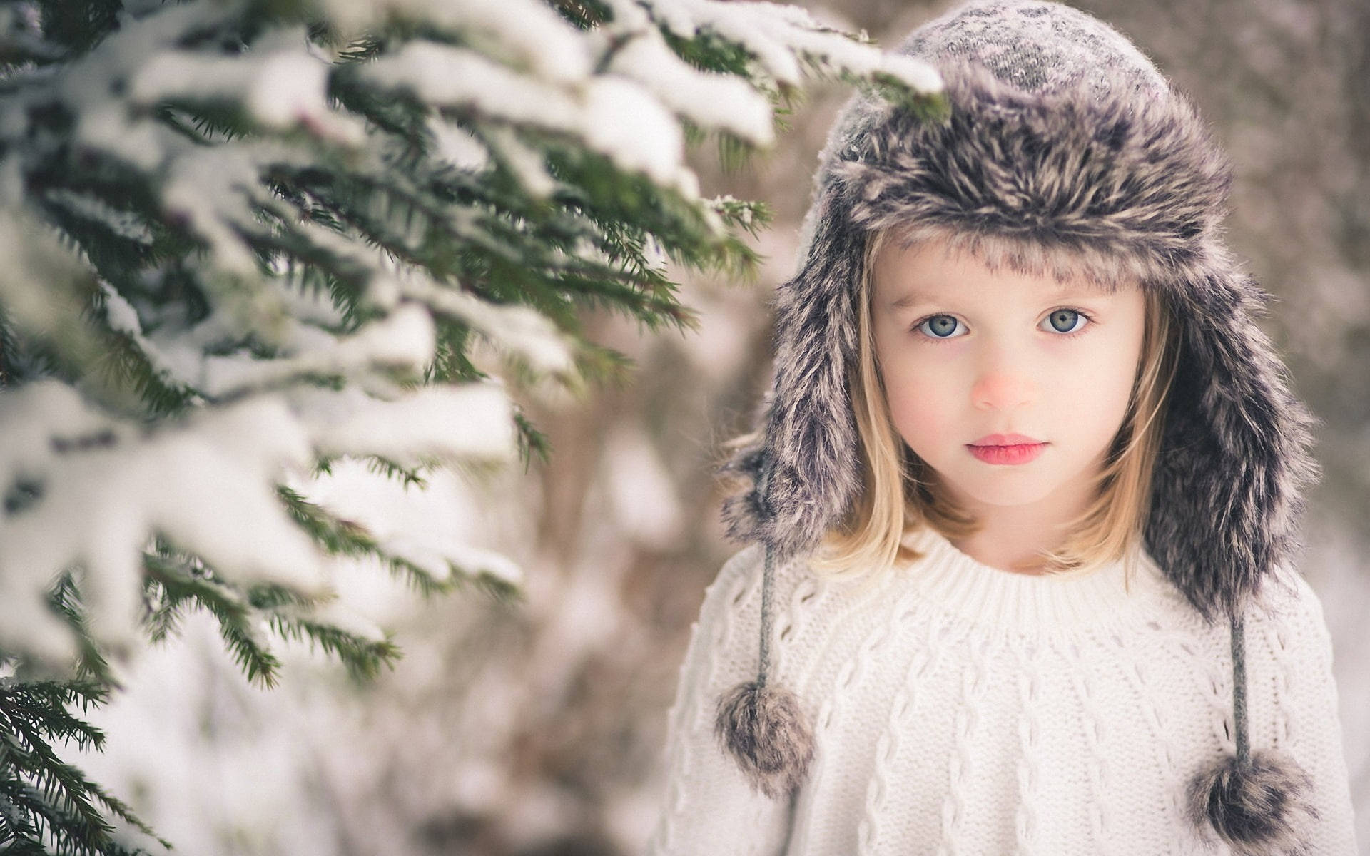 Cute Winter 桌布 壁紙 — 一個戴著毛帽、穿著白色針織毛衣的小女孩站在冬季森林中積雪的常綠樹枝旁。