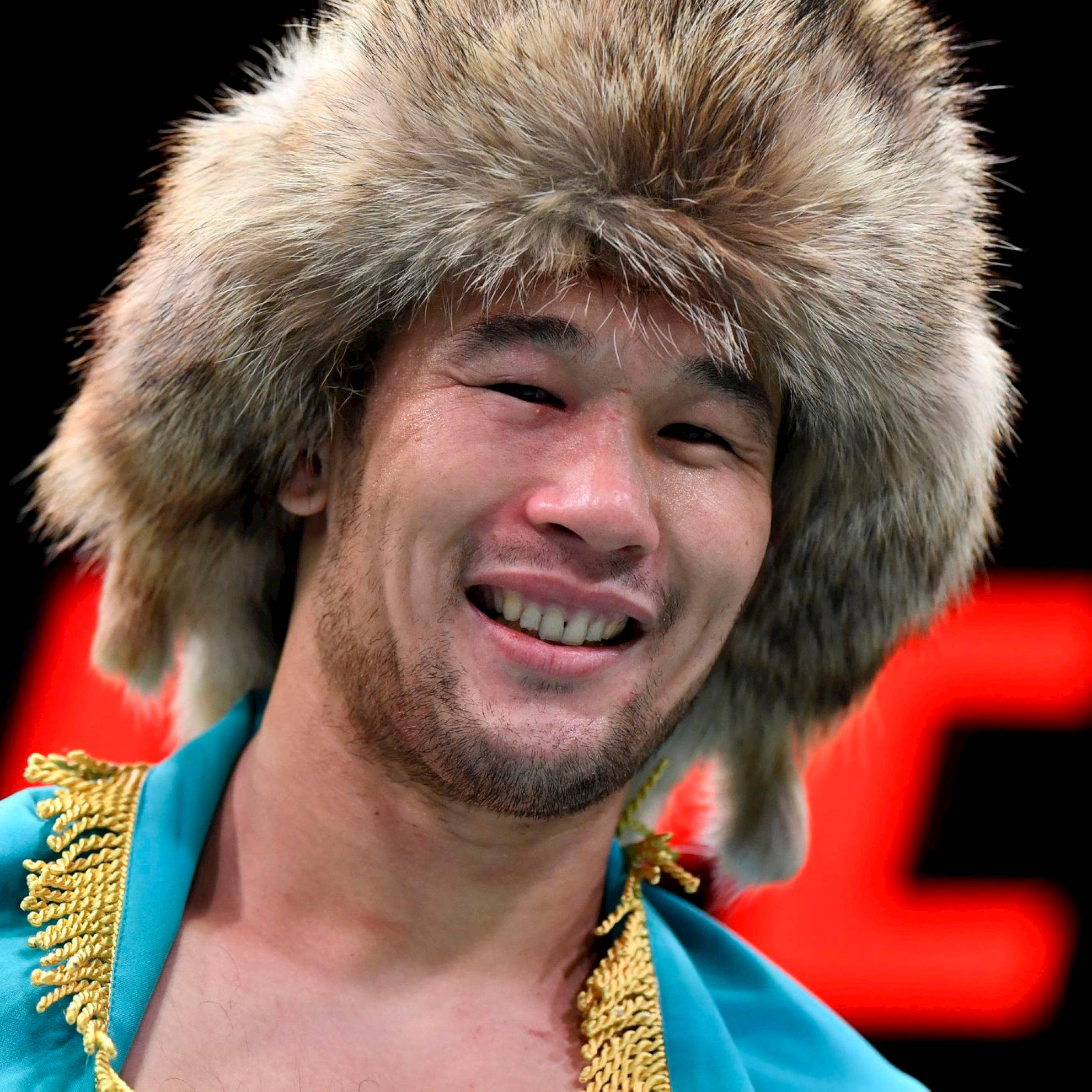 UFC 桌布 壁紙 — 一個戴著毛帽、身穿帶金色飾邊的綠松石色禮袍的男子，在紅色背景前微笑。