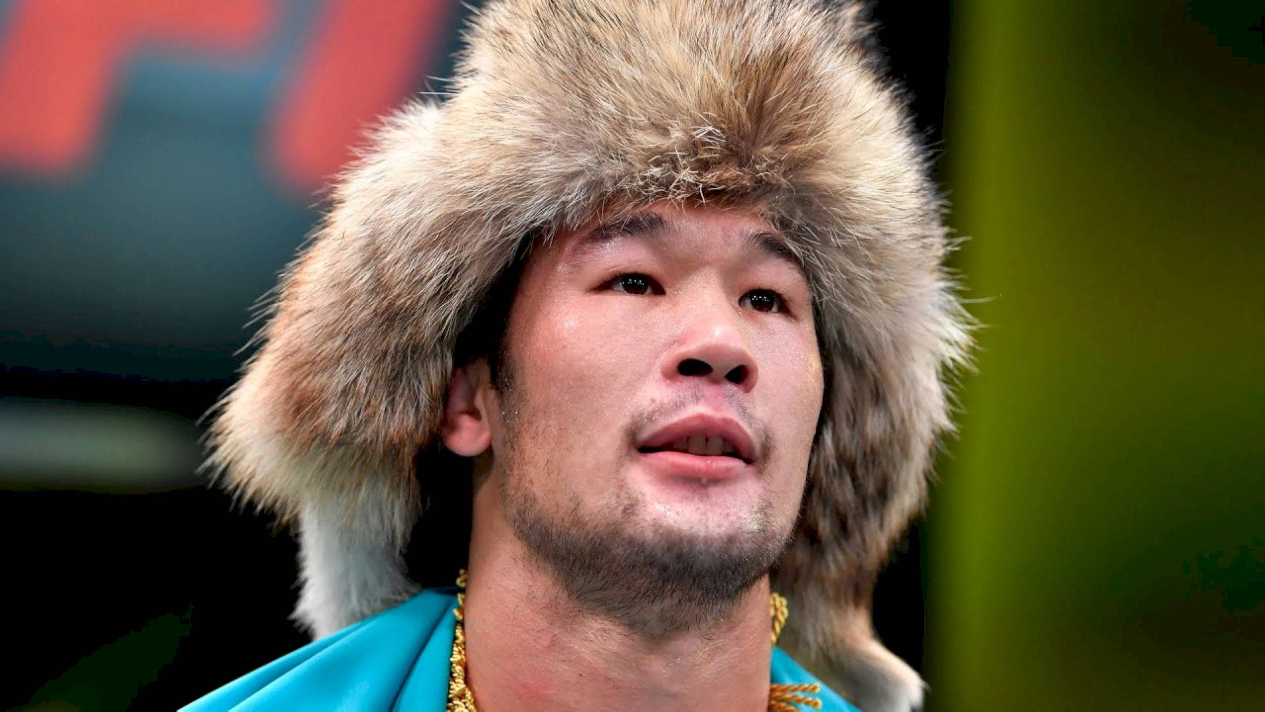UFC 桌布 壁紙 — 一個戴著毛帽和金鏈的男人，在模糊多彩的背景前，帶著專注的表情向上看。