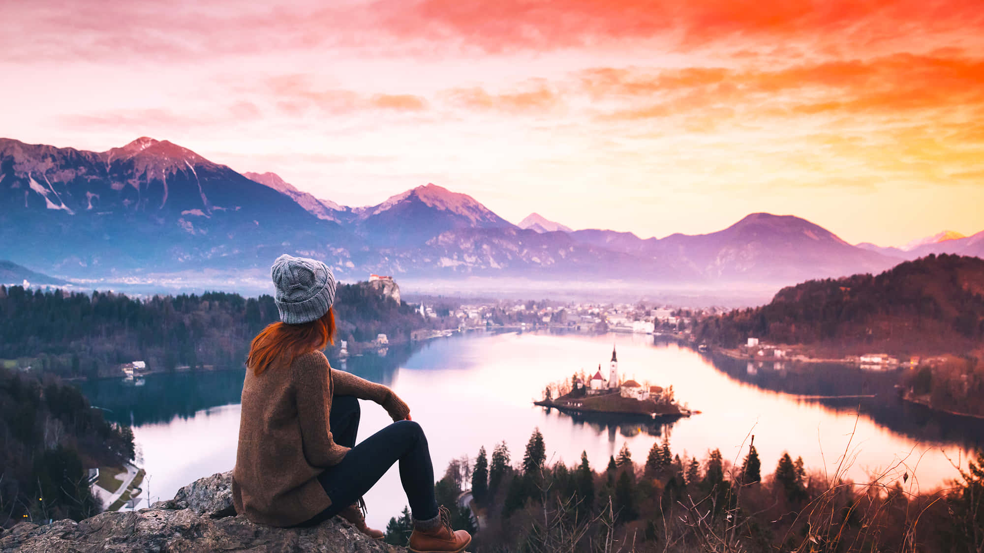 Travel Desktop 桌布 壁紙 — 一個戴著毛帽的人坐在懸崖上，俯瞰著一個寧靜的湖泊，湖中有教堂島和積雪的山峰，在充滿活力的日落天空下。