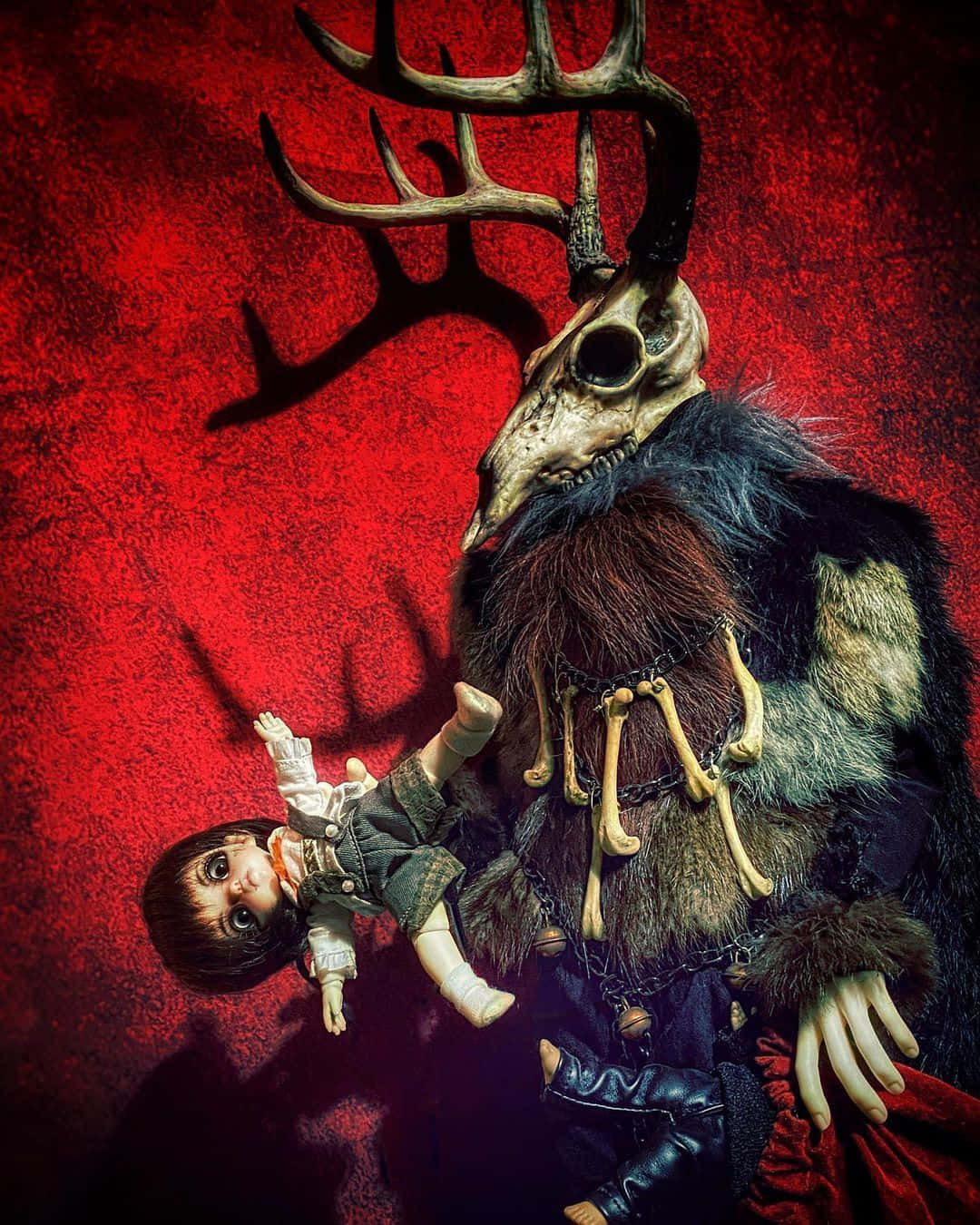 Krampus 桌布 壁紙 — 一個戴著毛皮和骨飾、頭部為鹿頭骨的威脅性人物，在紋理紅色的背景前抱著一個小玩偶。
