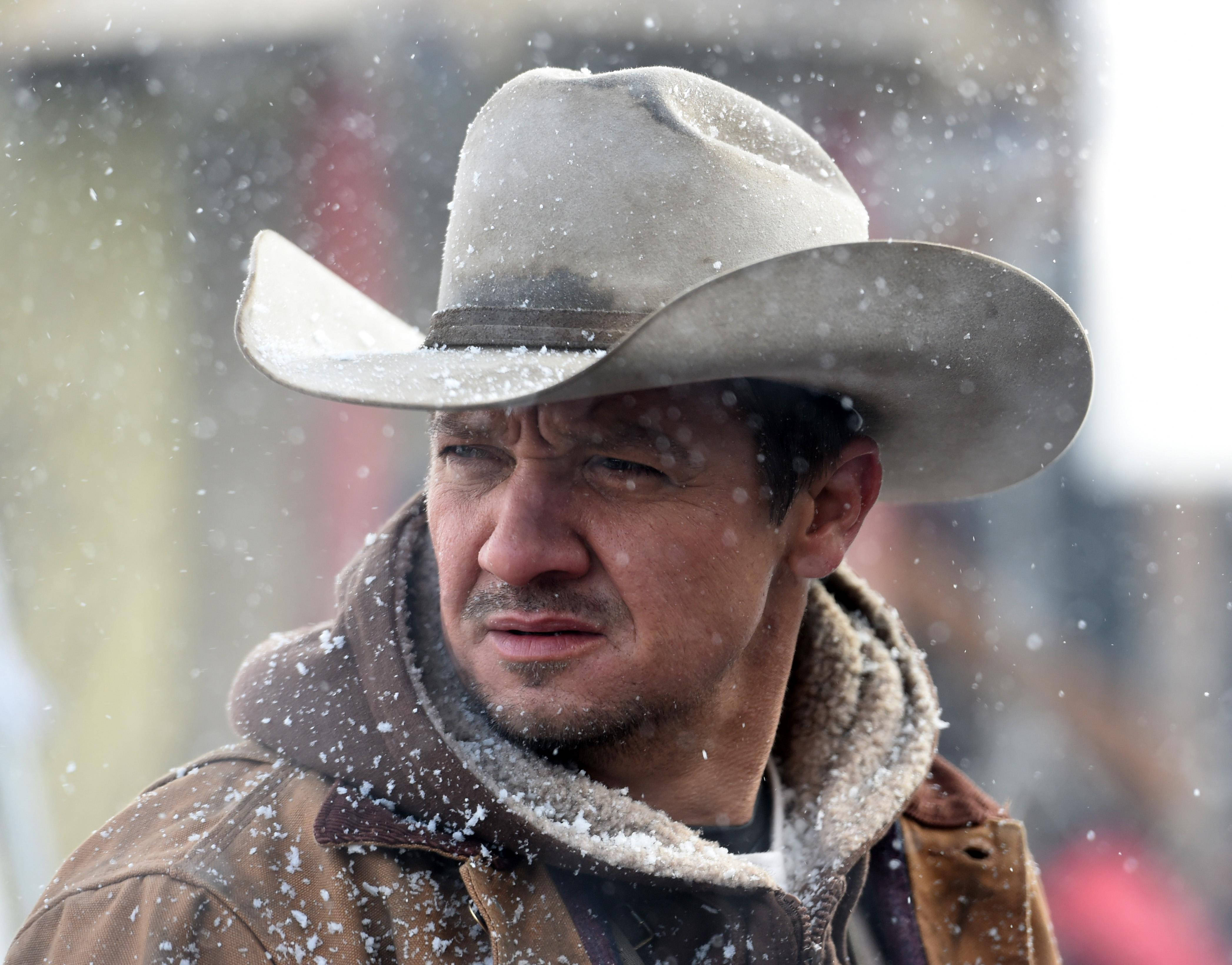 Jeremy Renner 桌布 壁紙 — 一個戴著沾滿雪花的牛仔帽、身穿外套的男人，在雪花飄落的冬日場景中肅穆站立。