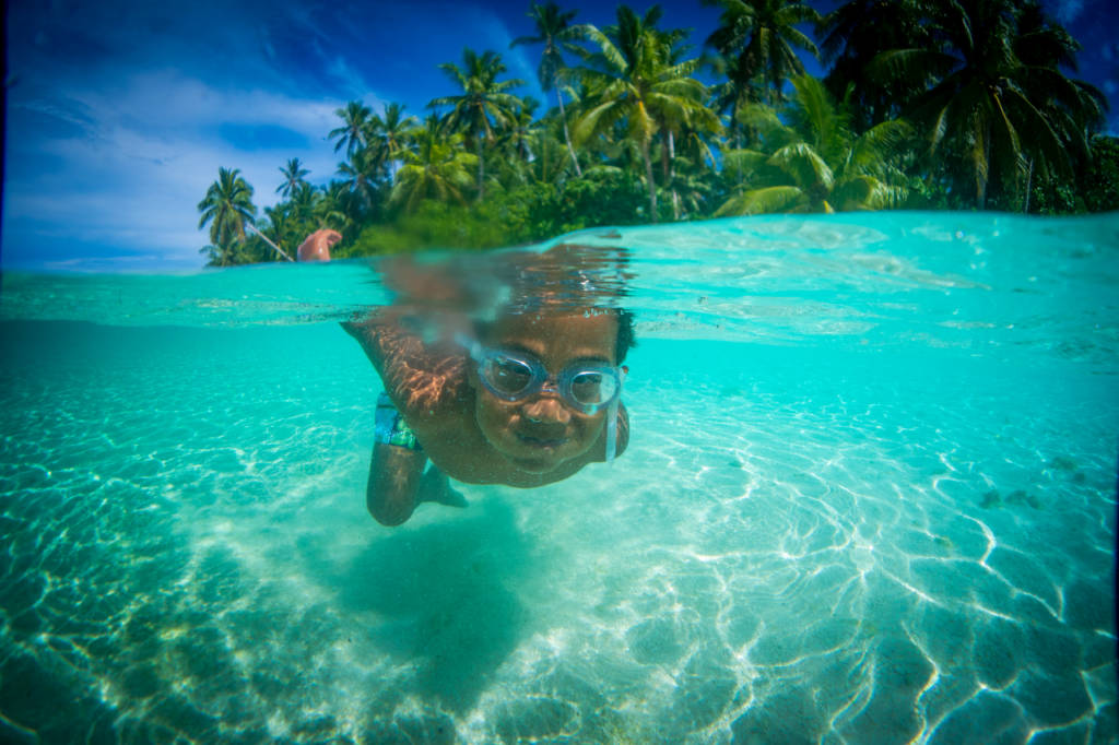 Swimming 桌布 壁紙 — 一個戴著泳鏡的小孩在水晶般清澈的碧綠水中游泳，上方可見棕櫚樹和藍天。