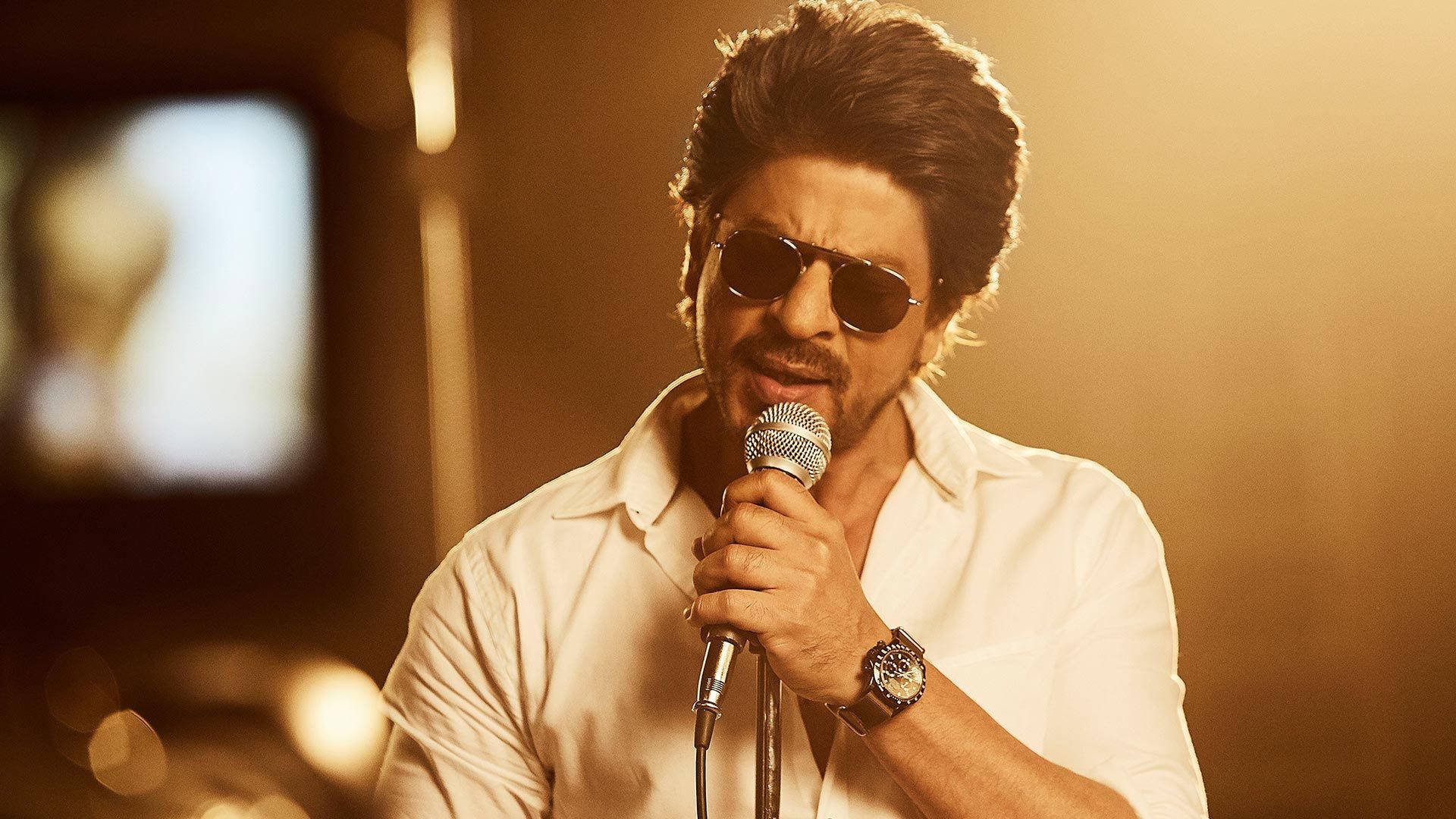 Shah Rukh Khan 桌布 壁紙 — 一個戴著深色太陽眼鏡、身穿白色襯衫的男人在溫暖的金色舞台燈光下對著麥克風唱歌。
