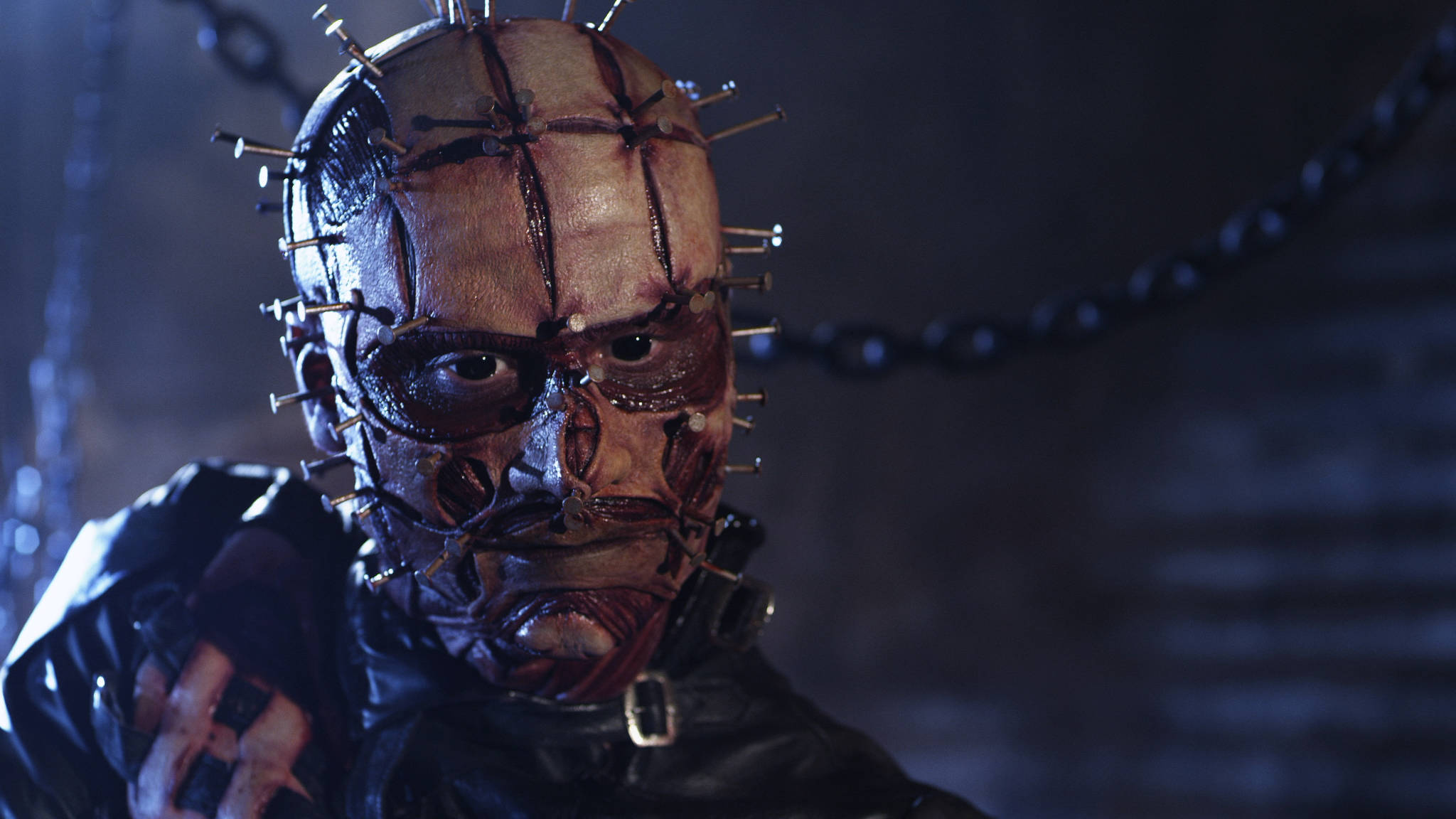 Pinhead 桌布 壁紙 — 一個戴著滿是針的面具的可怕人物，在昏暗且充滿鎖鏈的地牢中凝視著。