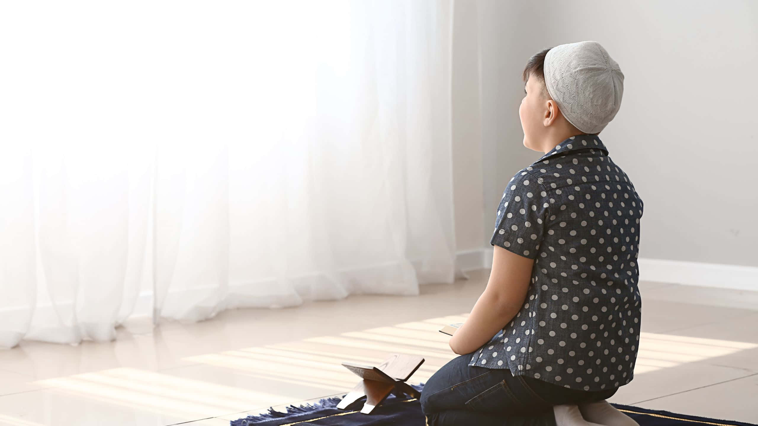 Praying 桌布 壁紙 — 一個戴著灰色基帕的小男孩在室內的祈禱毯上跪下，陽光透過他身後薄薄的白色窗簾照射進來。