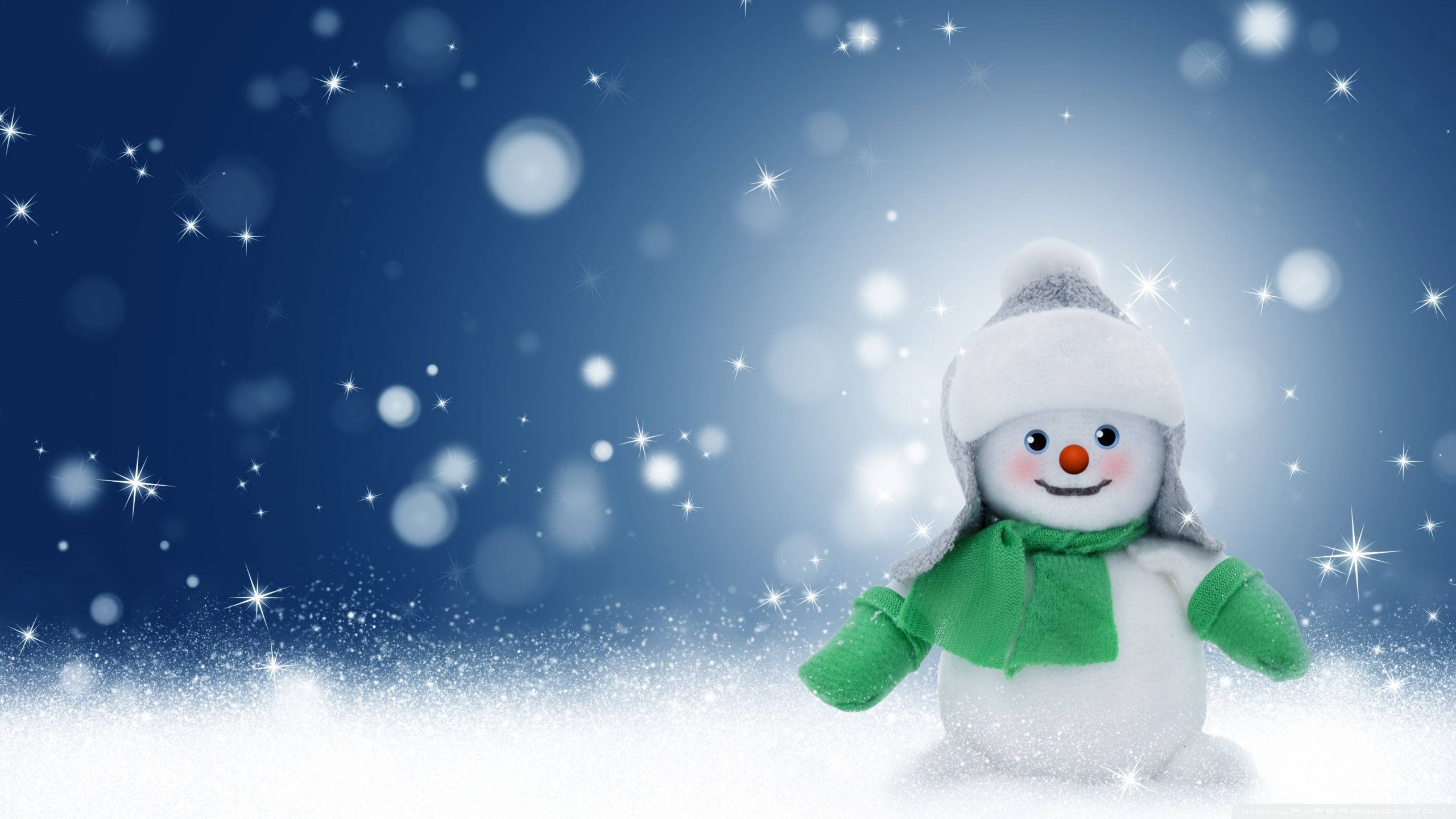Cute Snow 桌布 壁紙 — 一個戴著灰色帽子和綠色圍巾的快樂雪人，站在飄落的雪花中，背景是閃閃發光的藍色冬日天空。