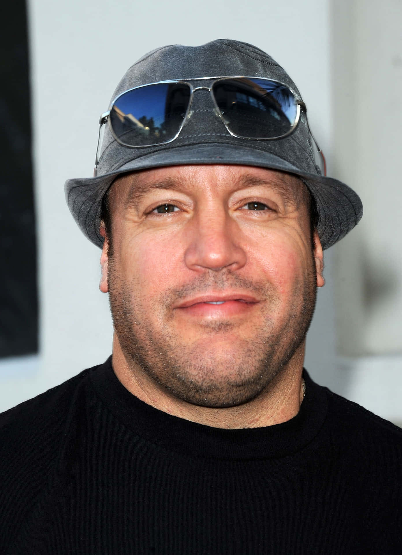 Kevin James 桌布 壁紙 — 一個戴著灰色漁夫帽、太陽眼鏡擱在頭上的男人，在近距離肖像照中對著鏡頭溫柔微笑。
