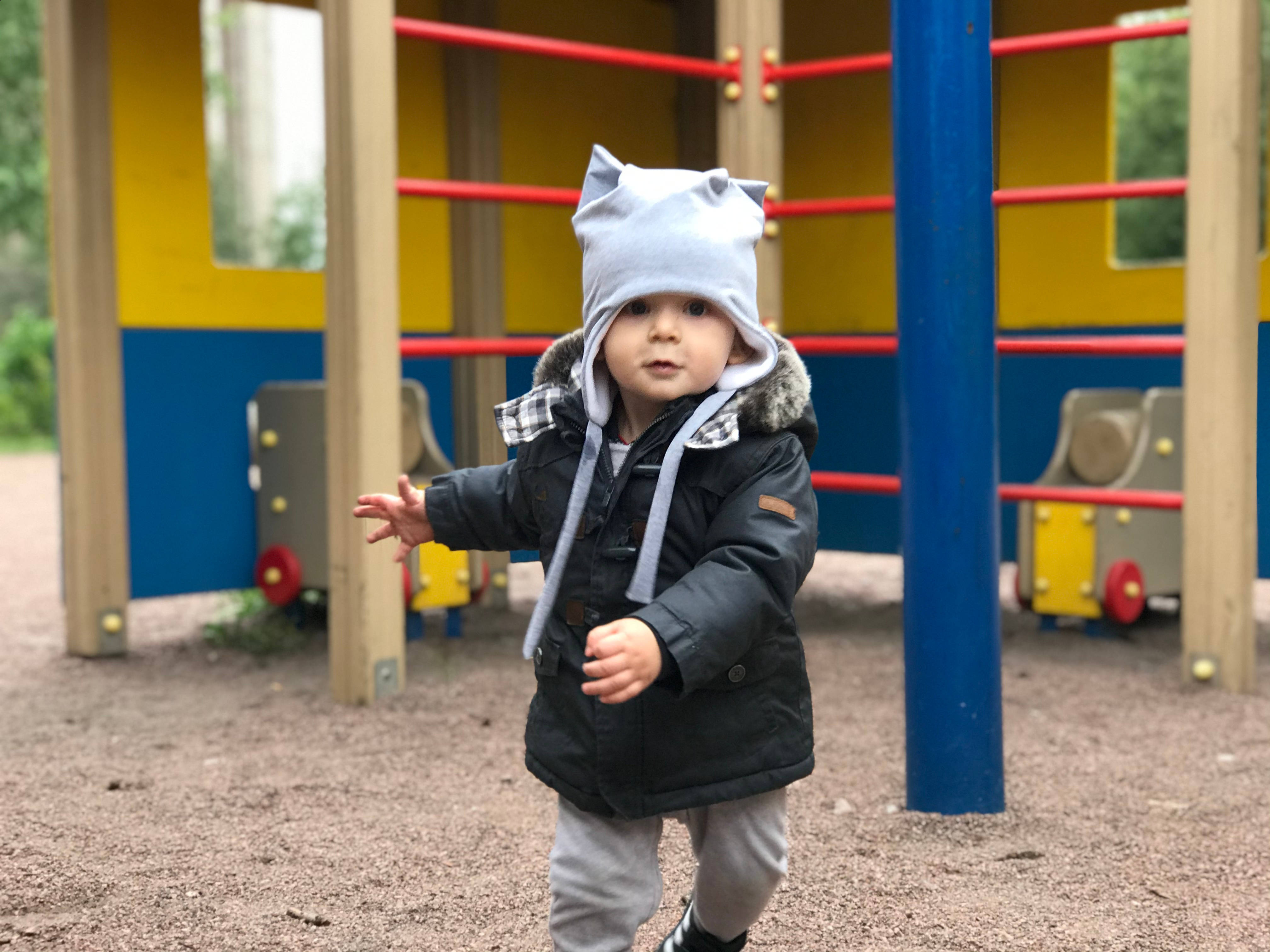 Playground 桌布 壁紙 — 一個戴著灰色貓耳帽和黑色冬季外套的幼兒站在遊樂場上，身後有色彩繽紛的遊樂設施。