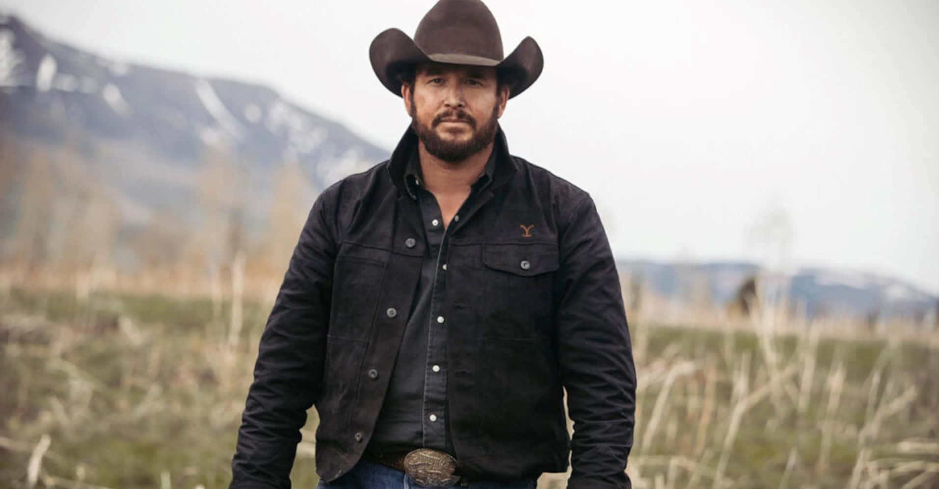 Yellowstone 桌布 壁紙 — 一個戴著牛仔帽、身穿深色外套的男人自信地站在草地上，背景是白雪覆蓋的山脈。