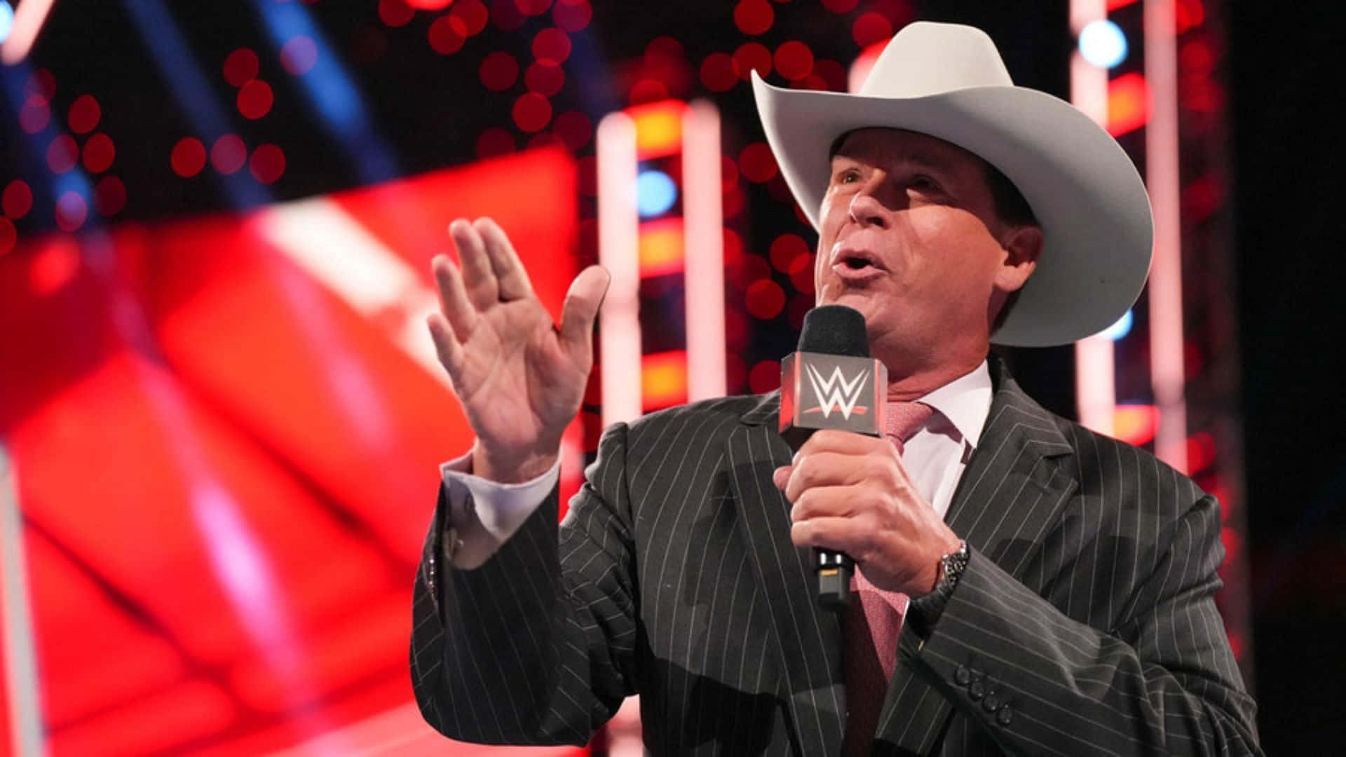 WWE 桌布 壁紙 — 一個戴著牛仔帽、身穿細條紋西裝的男人，在燈光明亮的舞台上熱情地比劃手勢，手握WWE麥克風。