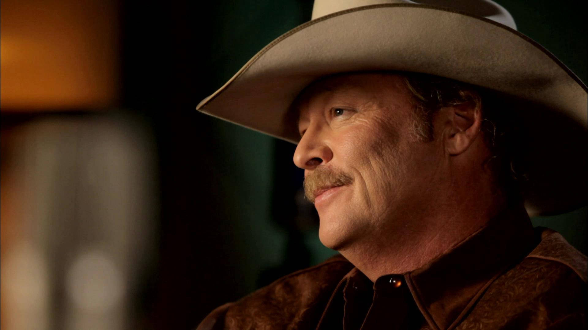 Alan Jackson 桌布 壁紙 — 一個戴著牛仔帽的男人在溫暖昏暗的燈光下若有所思地凝視，帶著一絲微笑。