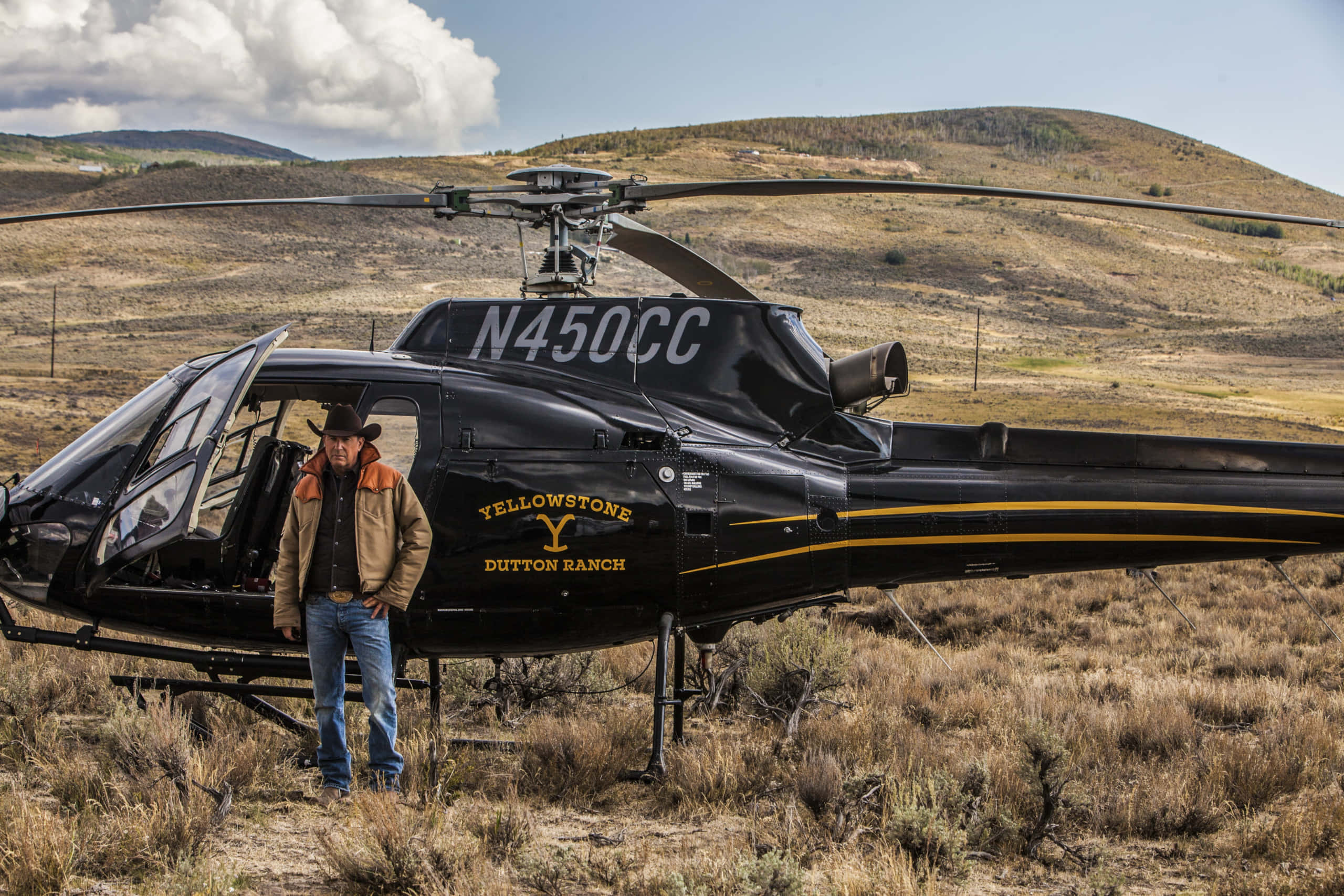 Yellowstone 桌布 壁紙 — 一個戴著牛仔帽的男人站在一架標有 'Yellowstone Dutton Ranch' 的黑色直升機旁邊，身處乾燥的草地上，天空部分多雲。
