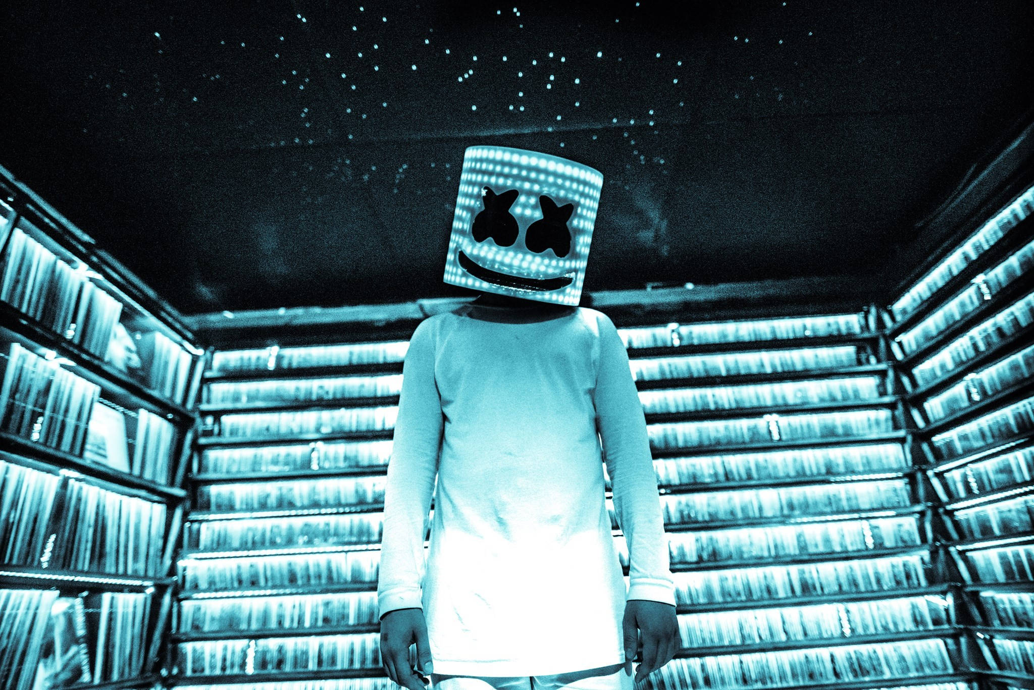 Marshmello 動態圖 桌布 壁紙 — 一個戴著發光棉花糖面具的人物站在一個有星星的房間裡，房間裡擺滿了發光的唱片架。