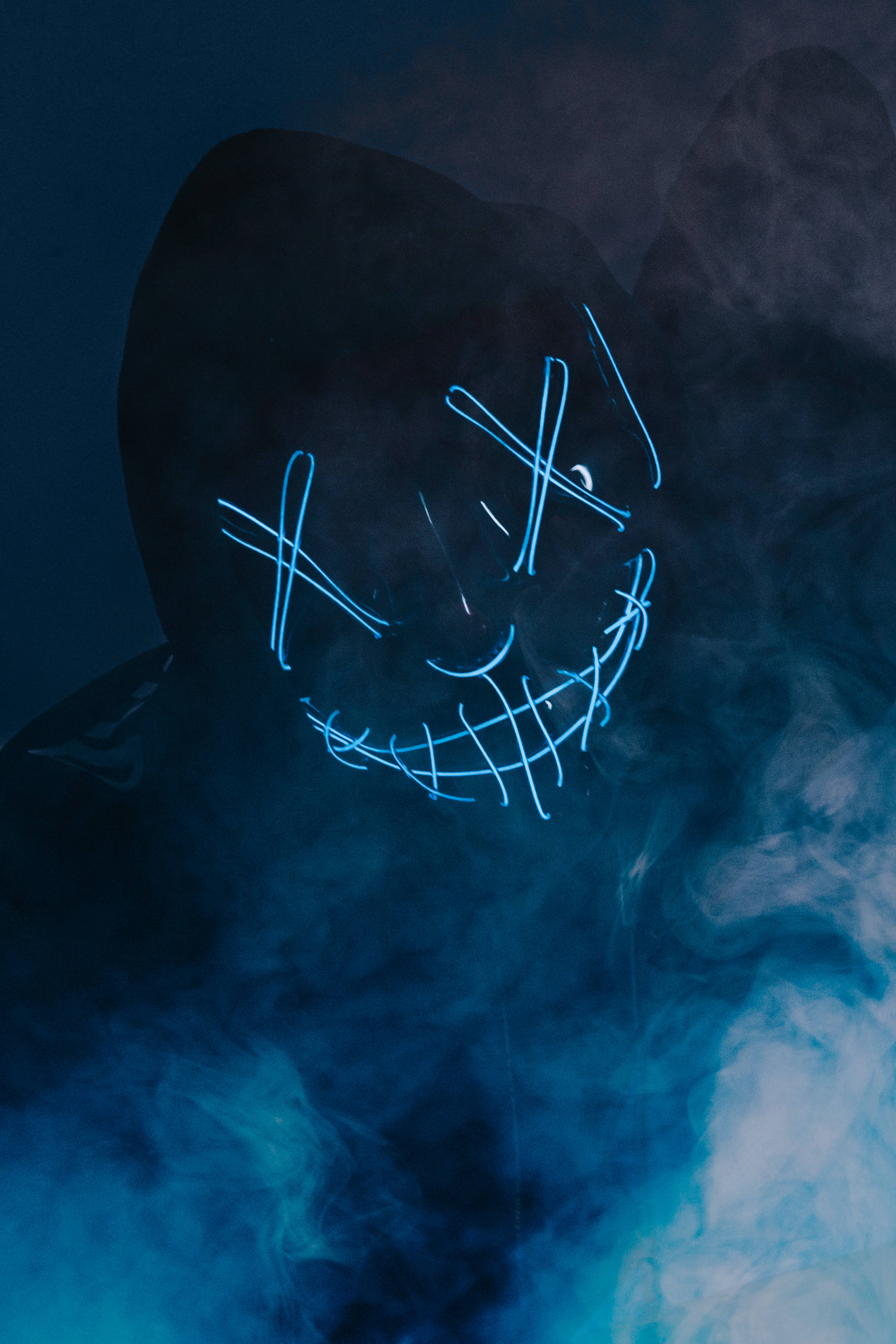 Smoke HD 桌布 壁紙 — 一個戴著發光藍色LED面具、帶有X眼睛和縫合笑容的兜帽人物，從深藍色迷霧中的旋轉煙霧中出現。