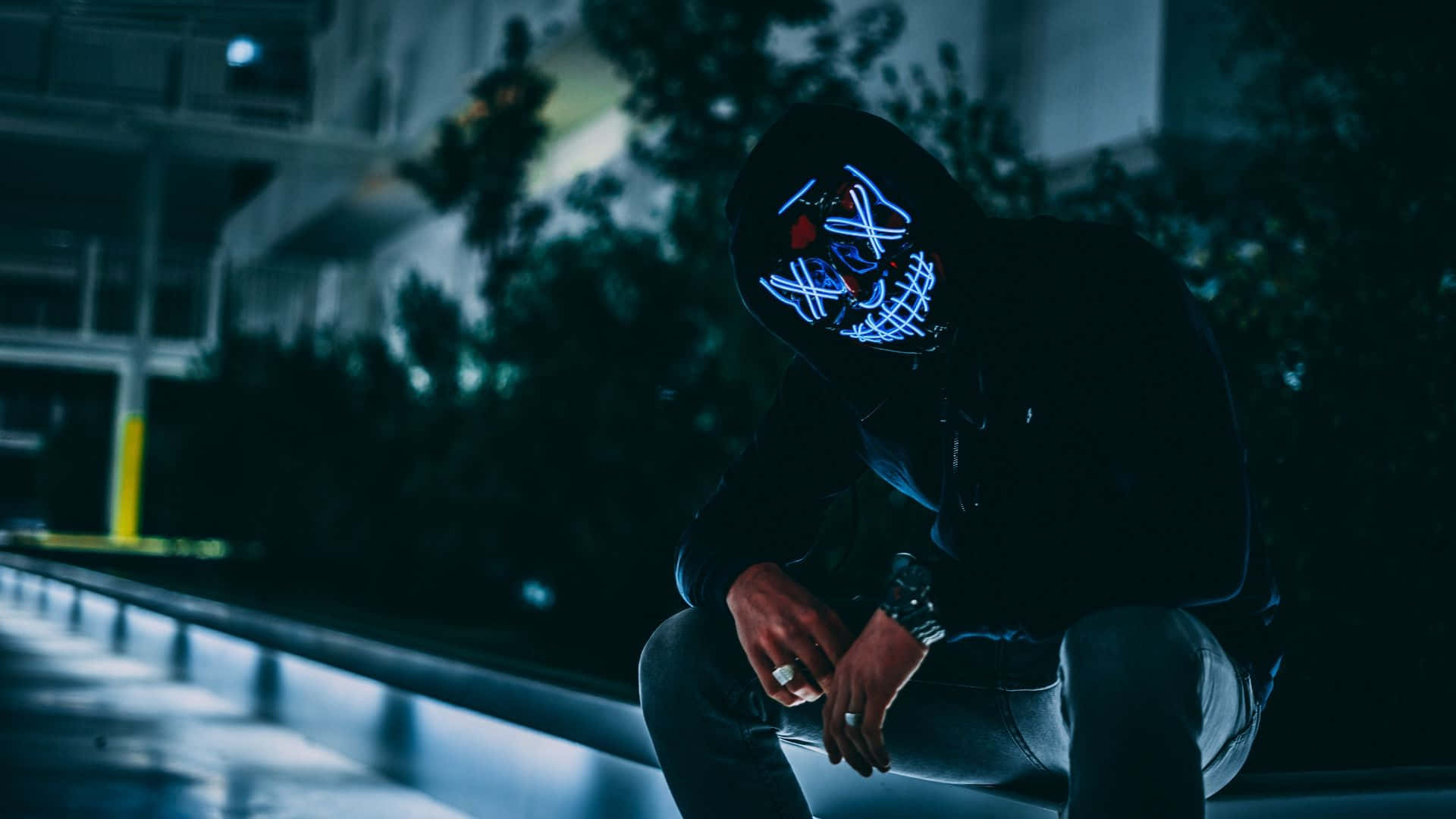 Mask 桌布 壁紙 — 一個戴著發光藍色LED面具的人，在夜晚昏暗的路燈下，獨自蹲在濕漉漉的城市邊緣。