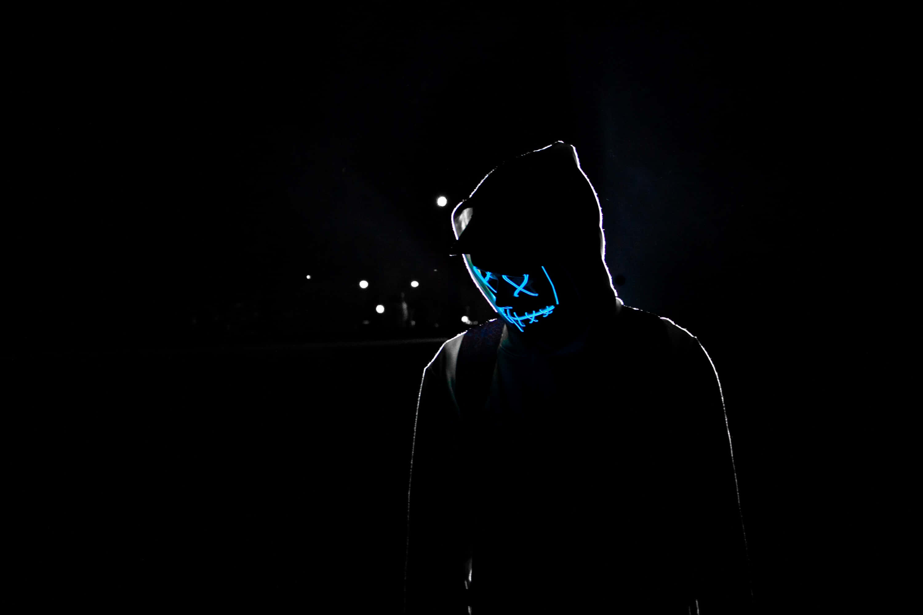 Neon Mask 桌布 壁紙 — 一個戴著發光藍色LED面具的兜帽人物在黑暗中，背景是夜晚遠處的城市景觀。