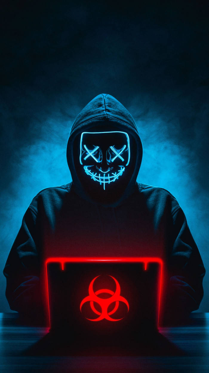 hacker 3d 桌布 壁紙 — 一個戴著發光藍色骷髏面具的兜帽人物坐在煙霧瀰漫的黑暗中，在顯示紅色生物危害符號的筆記型電腦上打字。