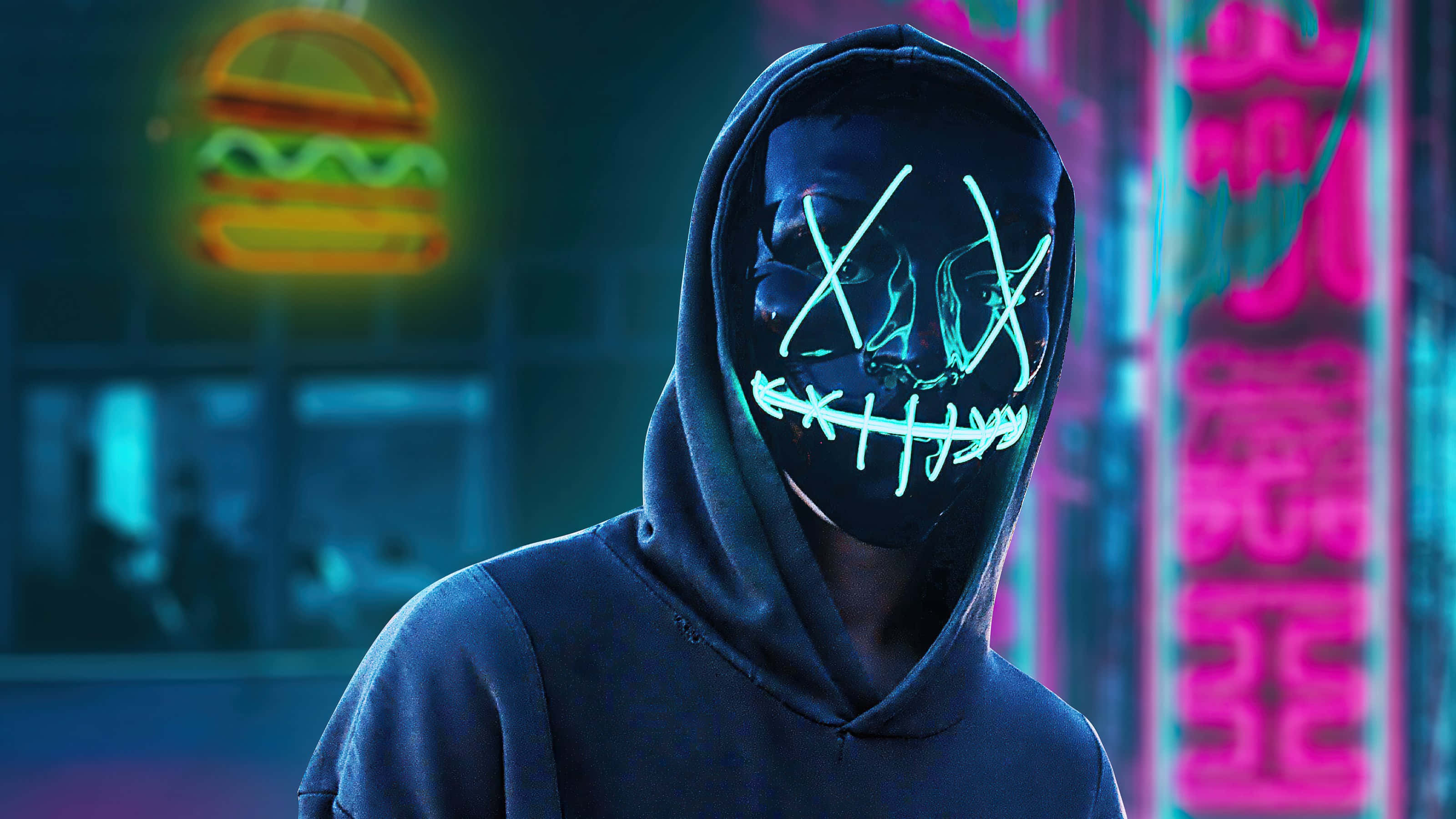 Mask Boy 桌布 壁紙 — 一個戴著發光霓虹面具的兜帽人物站在一條被鮮豔粉紅和藍色霓虹燈照亮的賽博朋克小巷中。