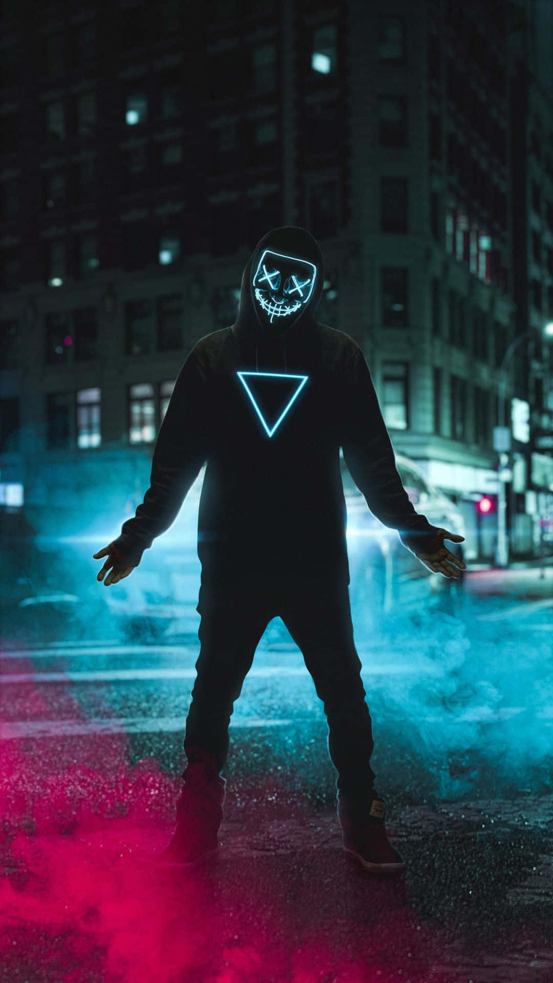 Mask 桌布 壁紙 — 一個戴著發光面具和連帽衫的人站在夜晚霓虹燈閃爍的都市街道上，紅色和藍色的煙霧在他們的腳邊旋轉。