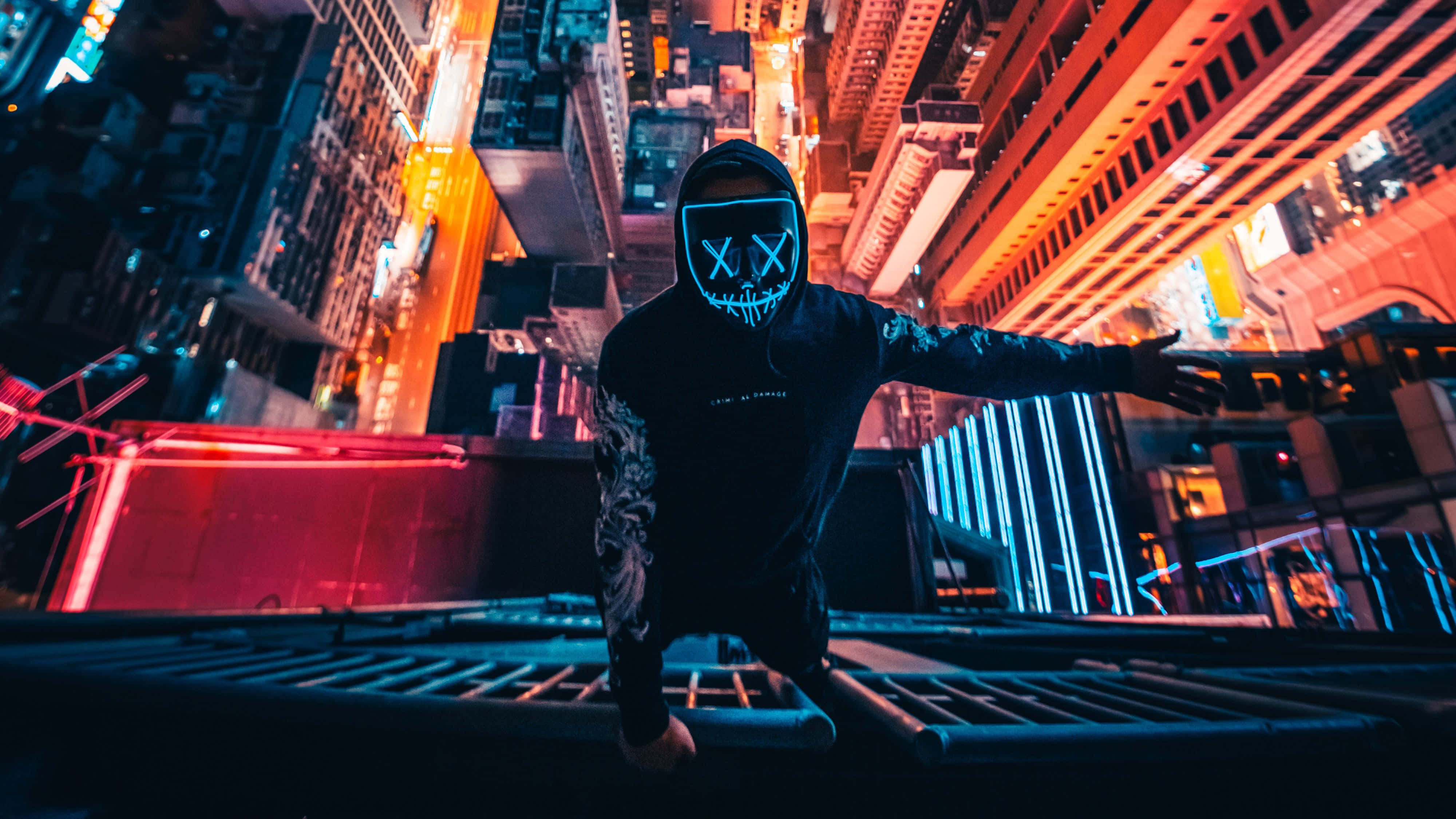 Neon Mask 桌布 壁紙 — 一個戴著發光面具的人蹲在屋頂上，背景是夜晚充滿活力的霓虹燈城市景觀。