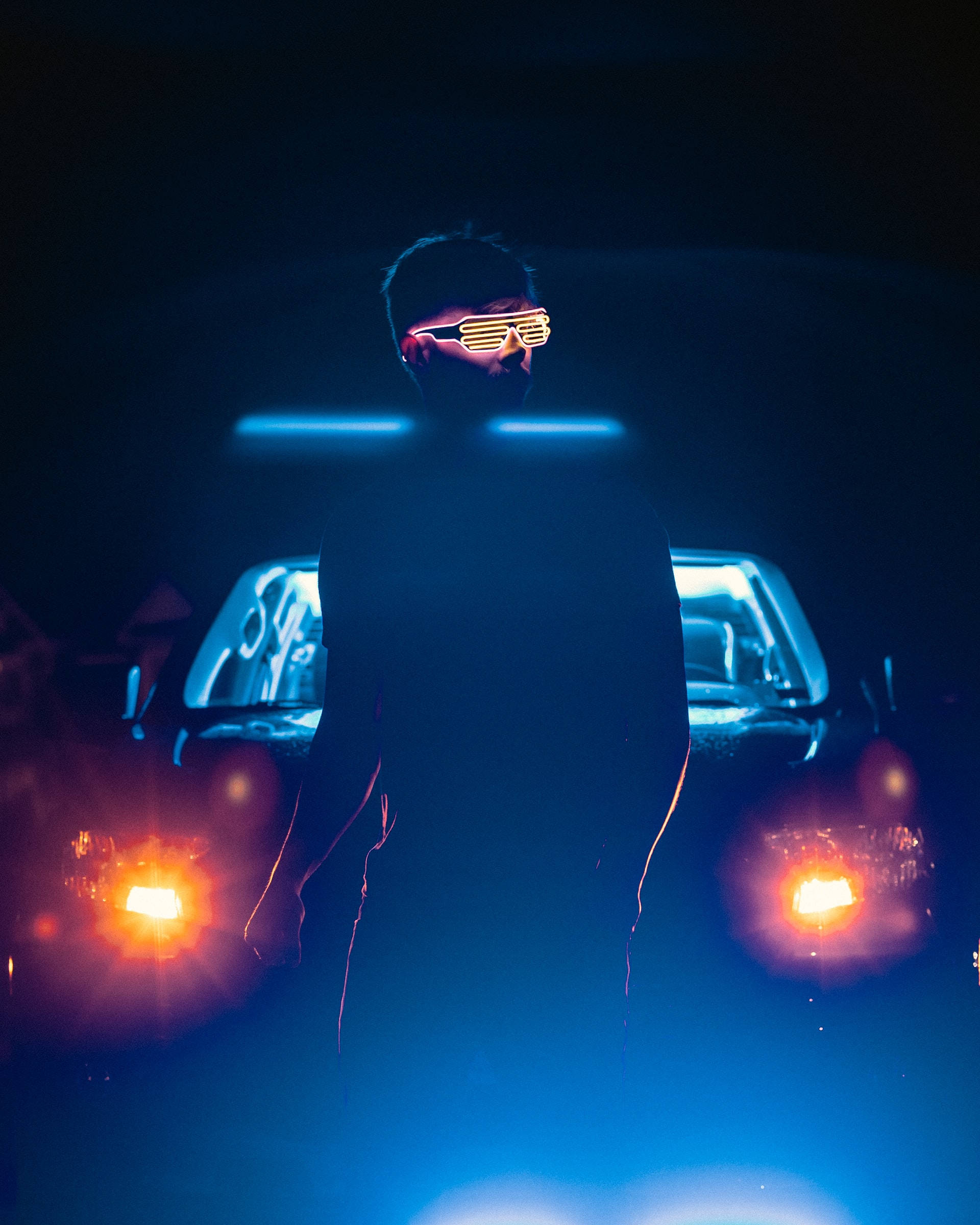Neon Lights Aesthetic 桌布 壁紙 — 一個戴著發光黃色百葉窗眼鏡的人的剪影，站在一輛帶有明亮橙色車頭燈的汽車前，場景為深藍色霓虹燈照明。