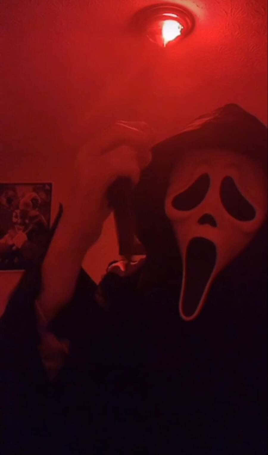Ghostface Aesthetic 桌布 壁紙 — 一個戴著白色Ghostface面具的人，在昏暗房間的單一紅色天花板燈下拿著一把刀。